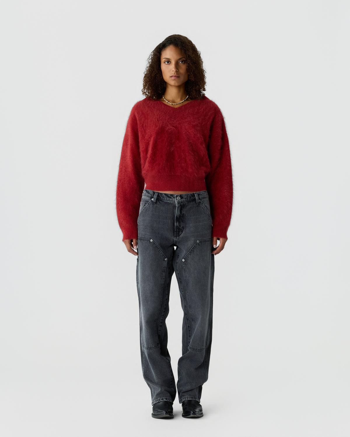 Woman with curly hair wearing a fuzzy red long-sleeve sweater and dark grey relaxed-fit jeans standing against a plain light background.