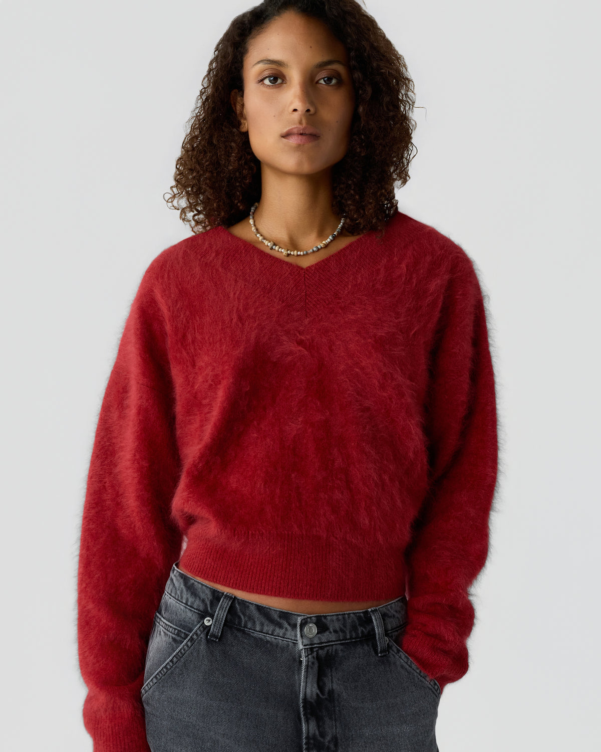 Woman wearing a red fuzzy sweater with a V-neck and long sleeves paired with black washed jeans, standing against a plain gray background.