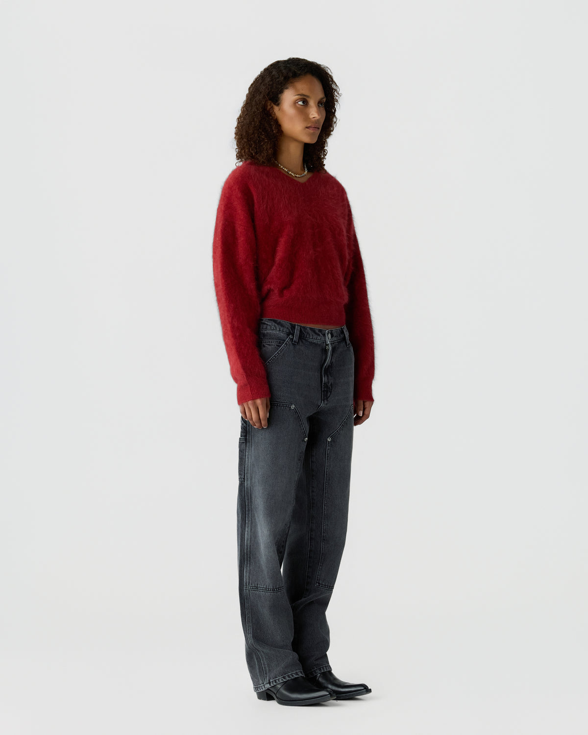 Woman with curly hair wearing a red fuzzy sweater, dark wide-legged jeans, and black pointed boots, standing against a plain white background.
