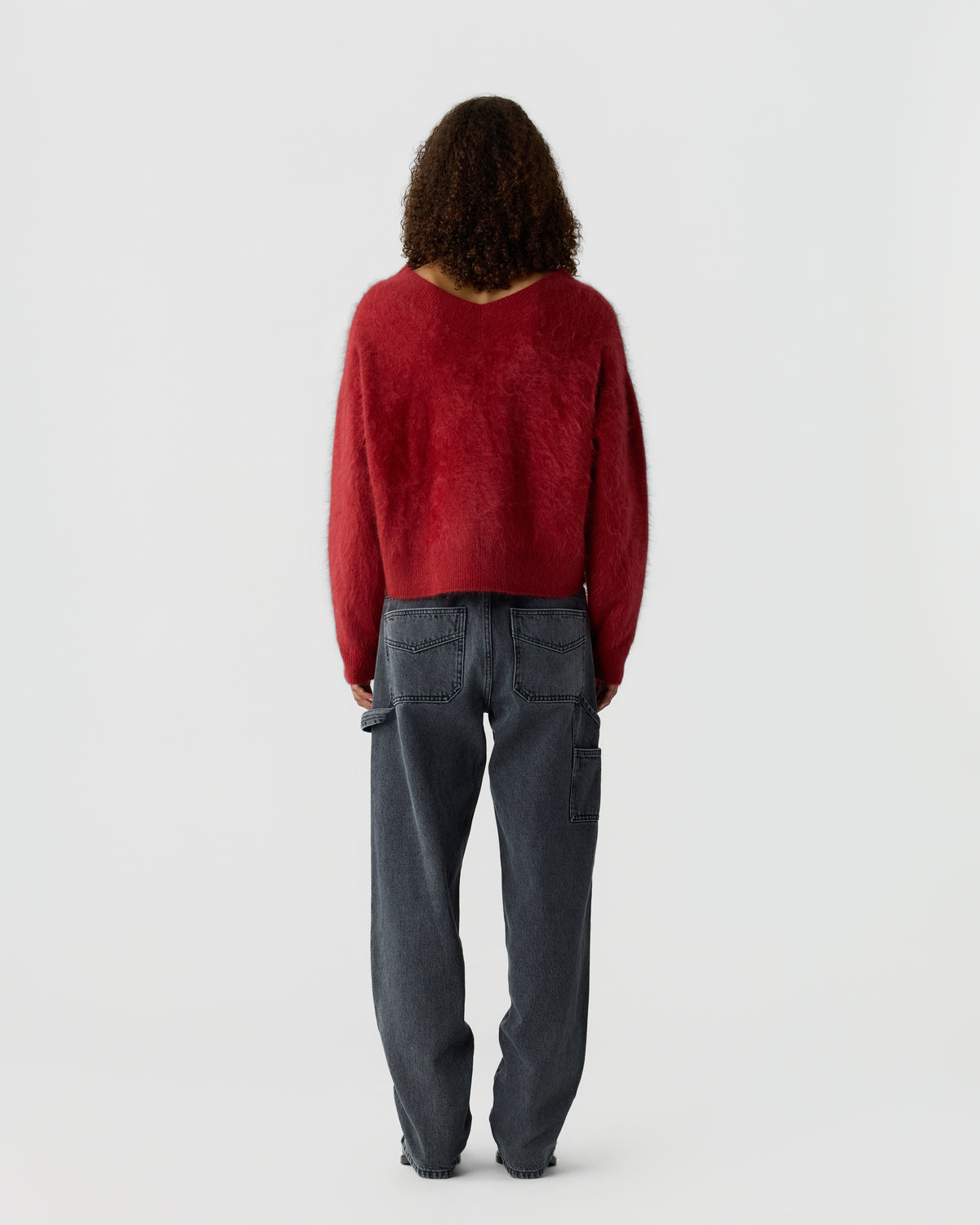 Person with curly hair standing facing away, wearing a red fuzzy long-sleeve sweater and loose-fitting dark gray pants against a plain white background.