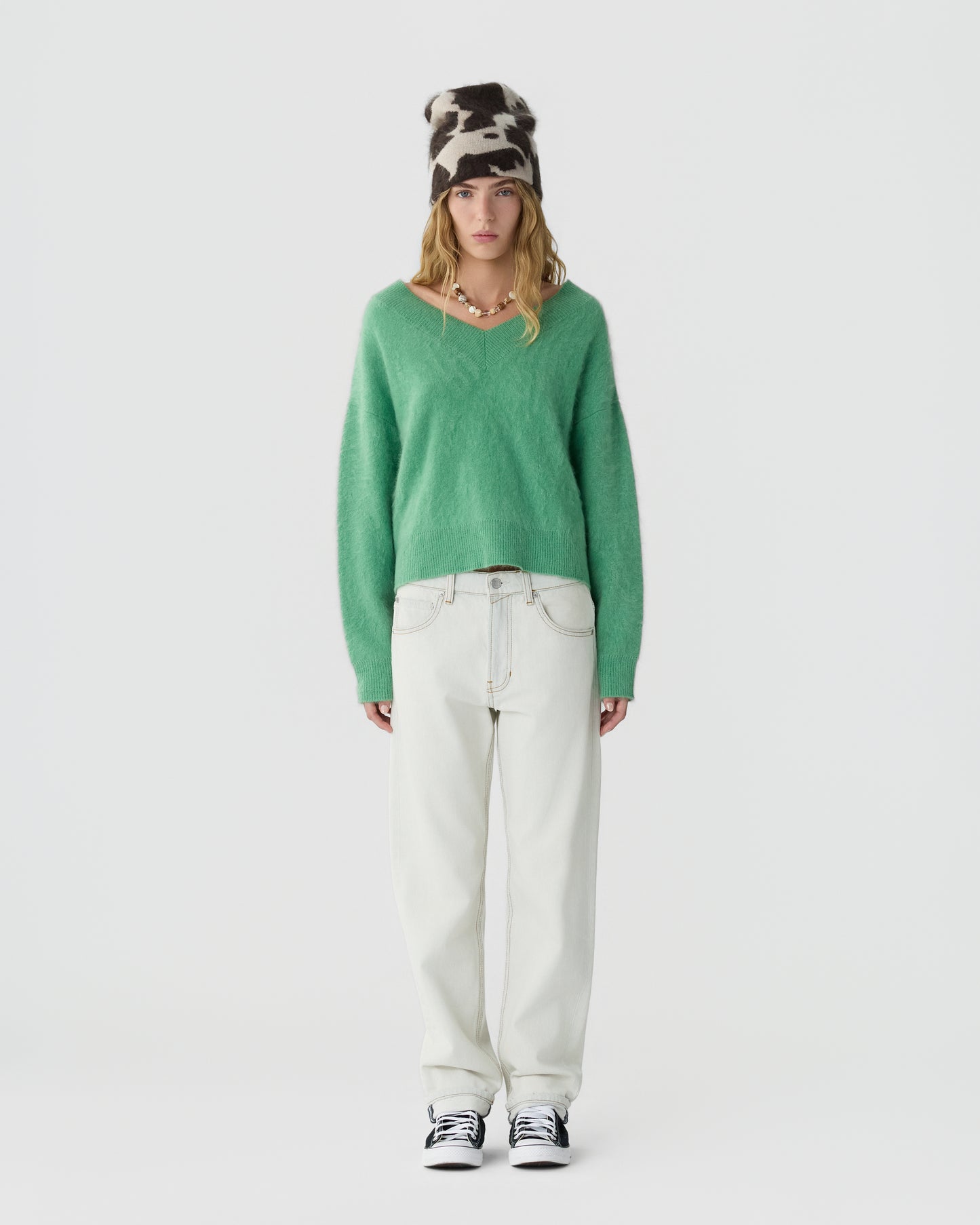 A person wearing a green fuzzy V-neck sweater, white high-waisted wide-leg jeans, black and white sneakers, and a black and white cow print beanie standing against a plain white background.