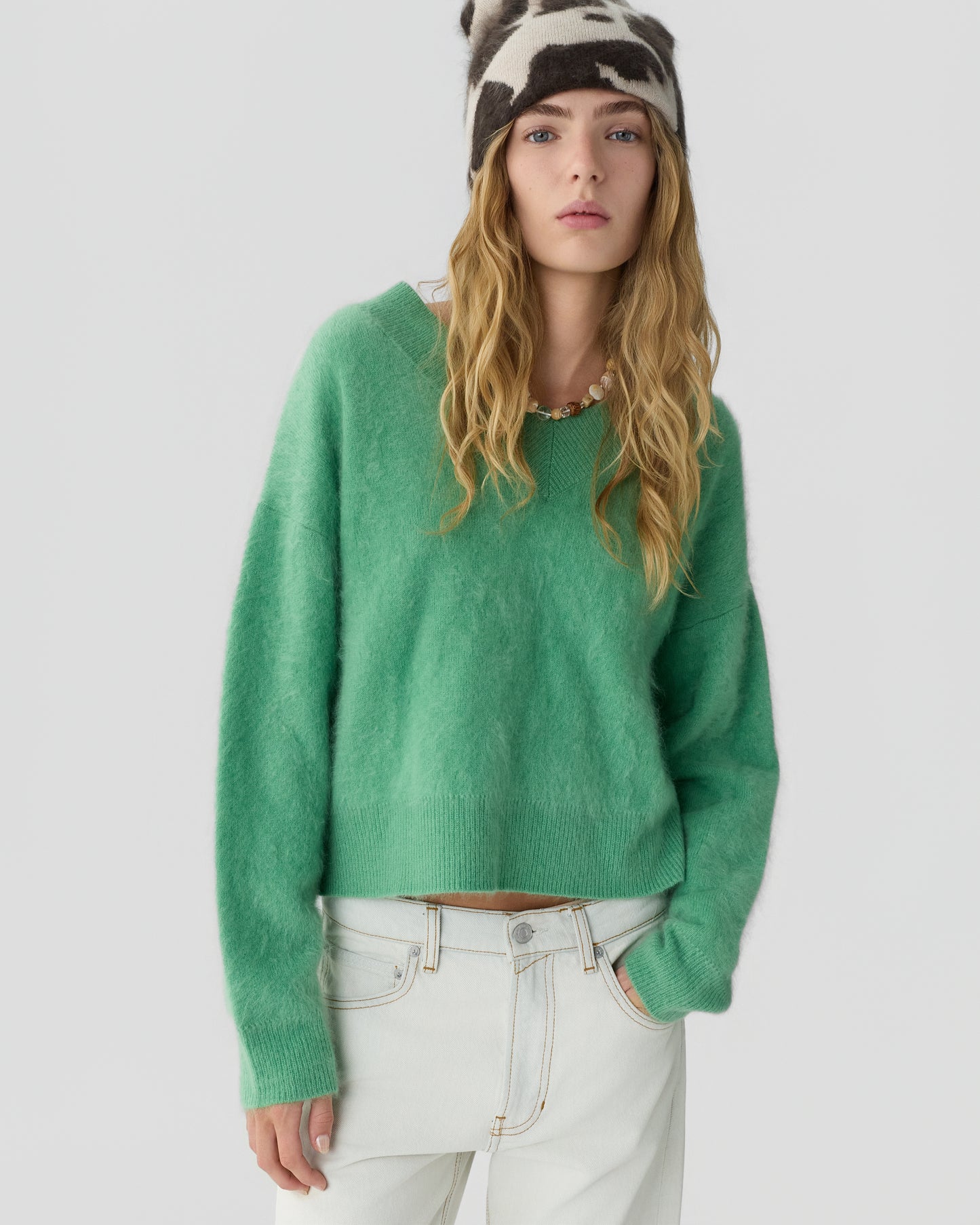 Young woman wearing a fuzzy green cashmere sweater, white high-waisted jeans, and a black-and-white patterned knit beanie standing against a plain light gray background.