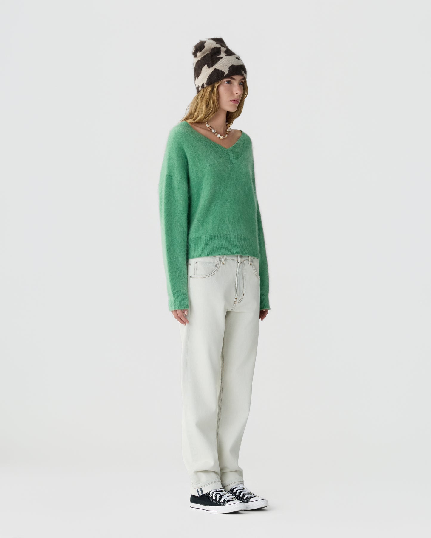 Young woman wearing a green V-neck sweater, white loose-fitting pants, black and white sneakers, and a black and white patterned knitted beanie on a plain light gray background.