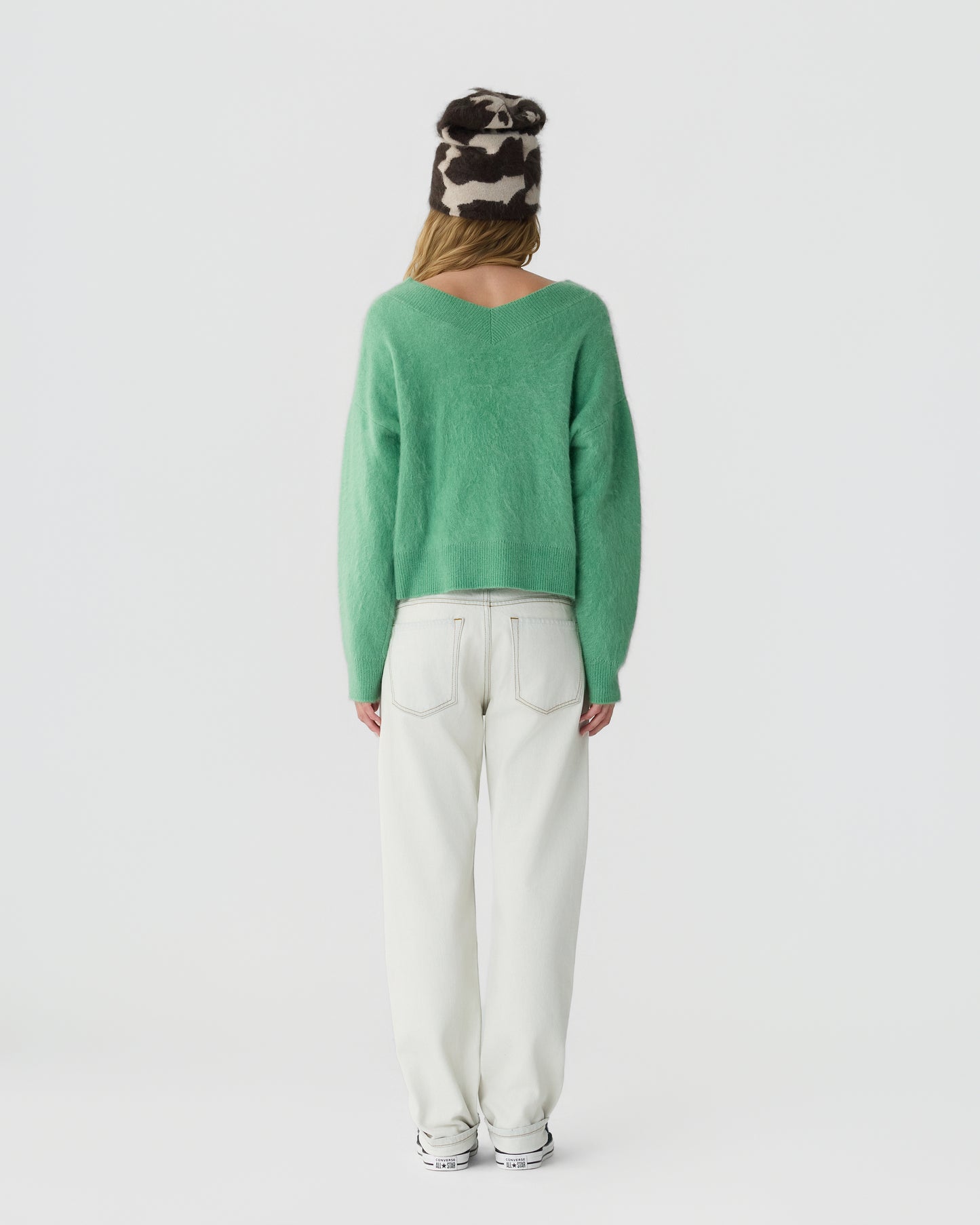 Woman standing facing away wearing a green fuzzy sweater, white high-waisted pants, white sneakers, and a black and white patterned beanie hat against a plain background.