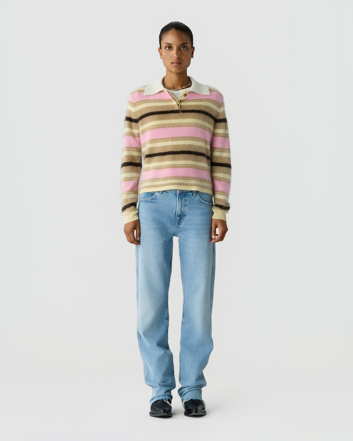 Model wearing a pink, cream, and brown striped collared sweater with light blue straight leg jeans and black shoes against a pale gray background