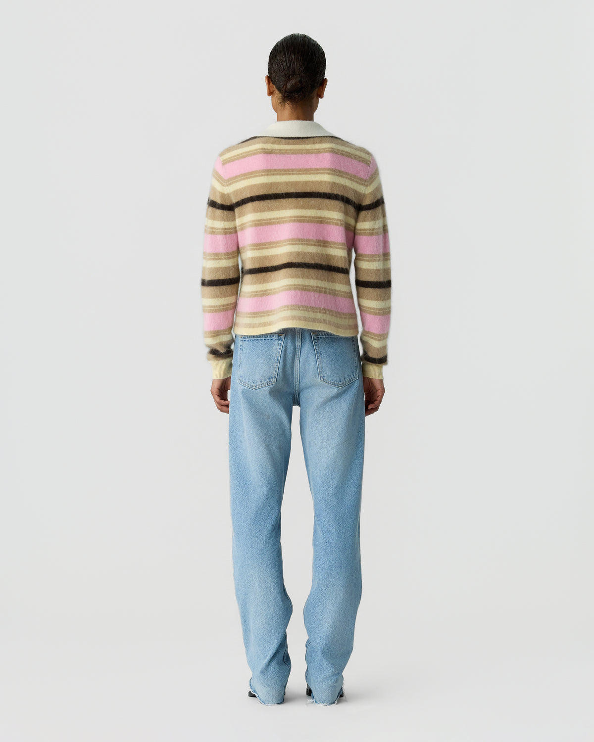 Back view of a person wearing a striped sweater in pink beige and black with light blue jeans against a pale gray background