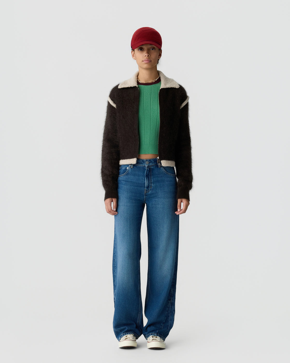 Full length portrait of a person wearing a red cap a dark shearling collar jacket green sweater blue wide leg jeans and white sneakers against a light gray background