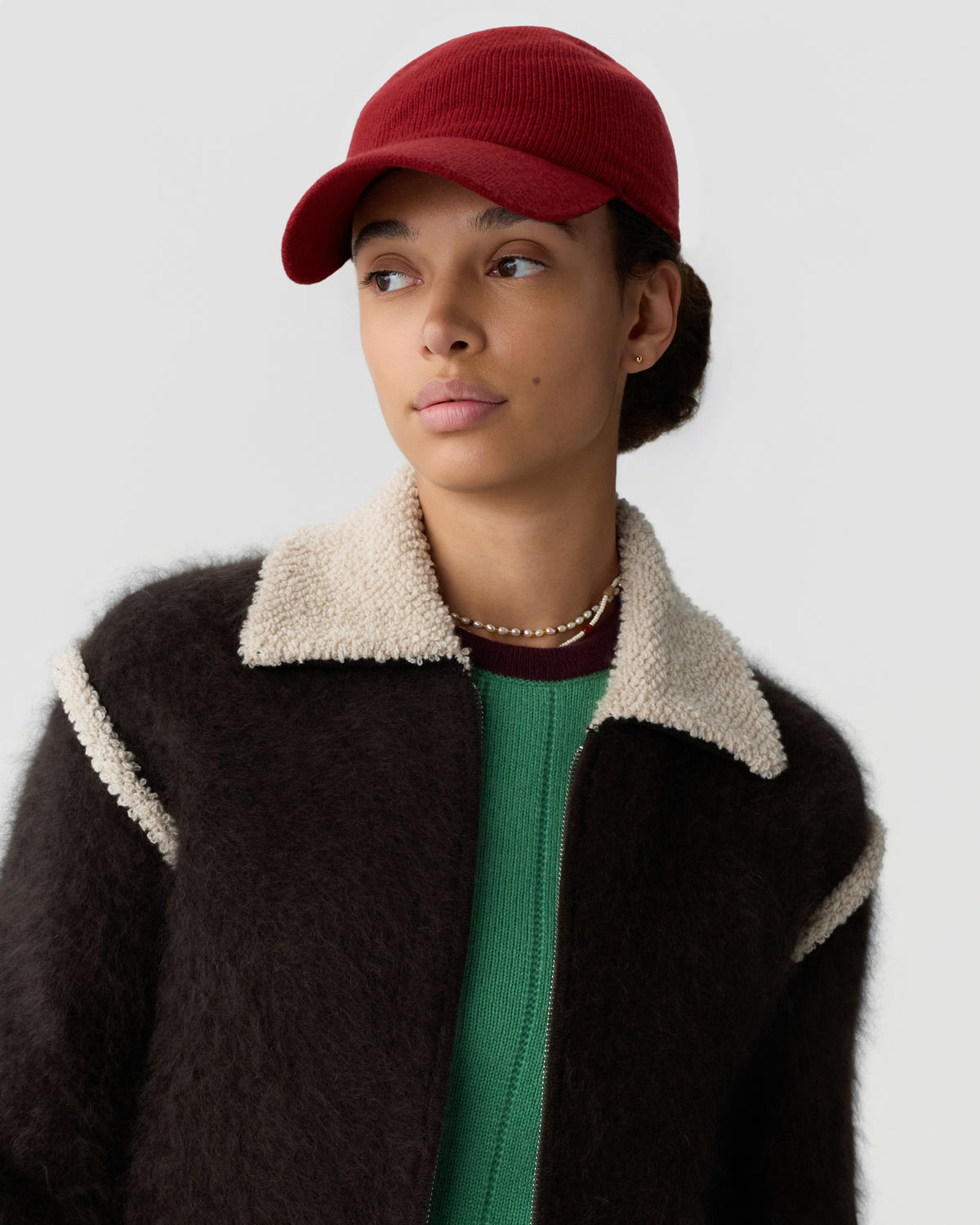 Woman wearing a red knit cap, black shearling jacket with a cream collar, and green sweater, looking to the side against a light gray background