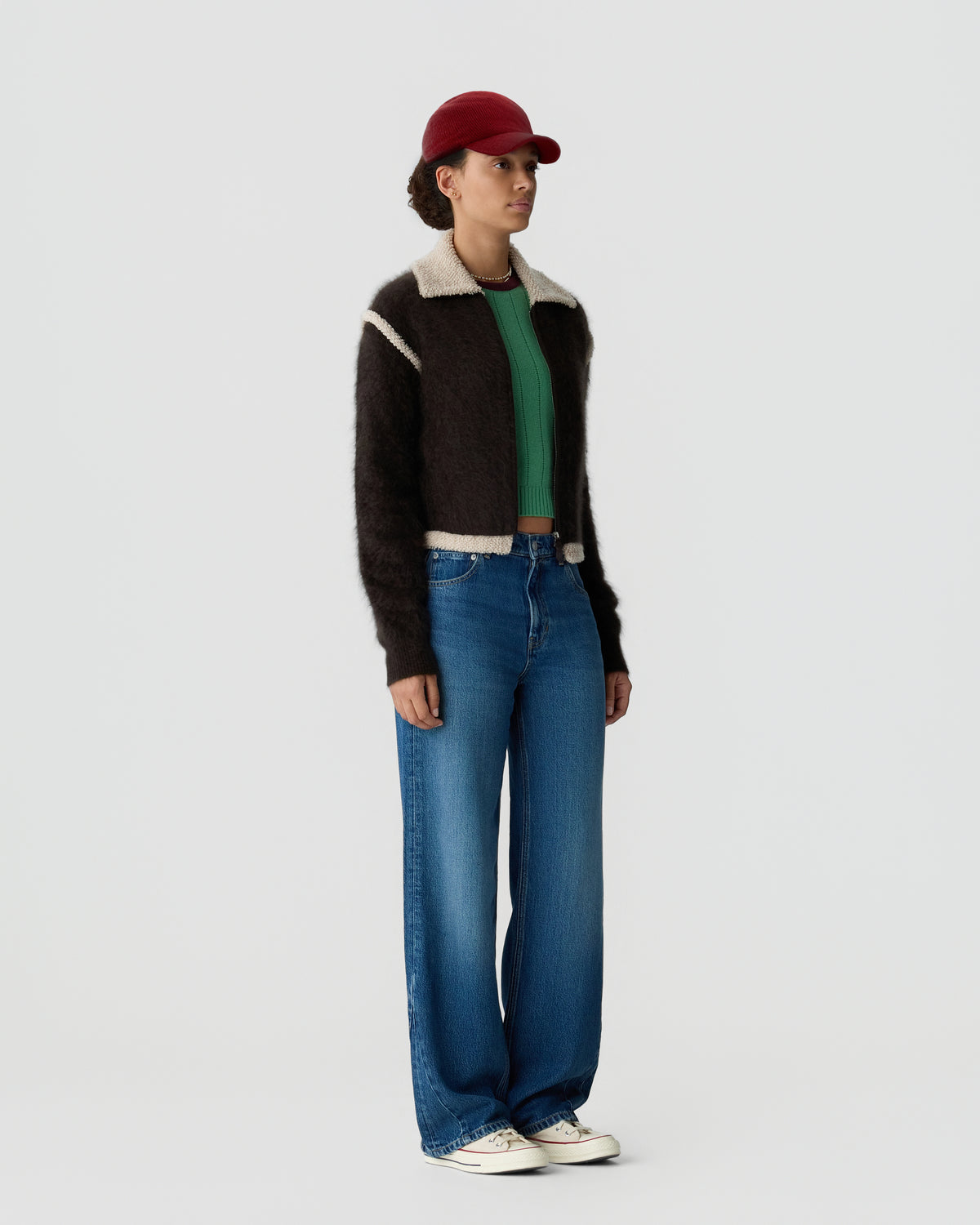 Full-length view of a person wearing a red cap brown collared jacket green sweater blue wide-leg jeans and white sneakers against a light background