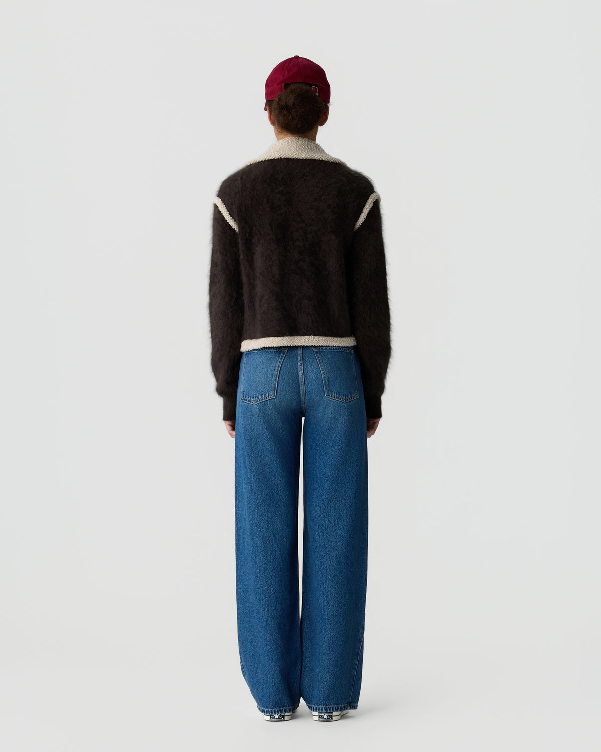 Back view of a person wearing a black knit sweater with cream trim, a red cap, and relaxed blue jeans against a light gray background