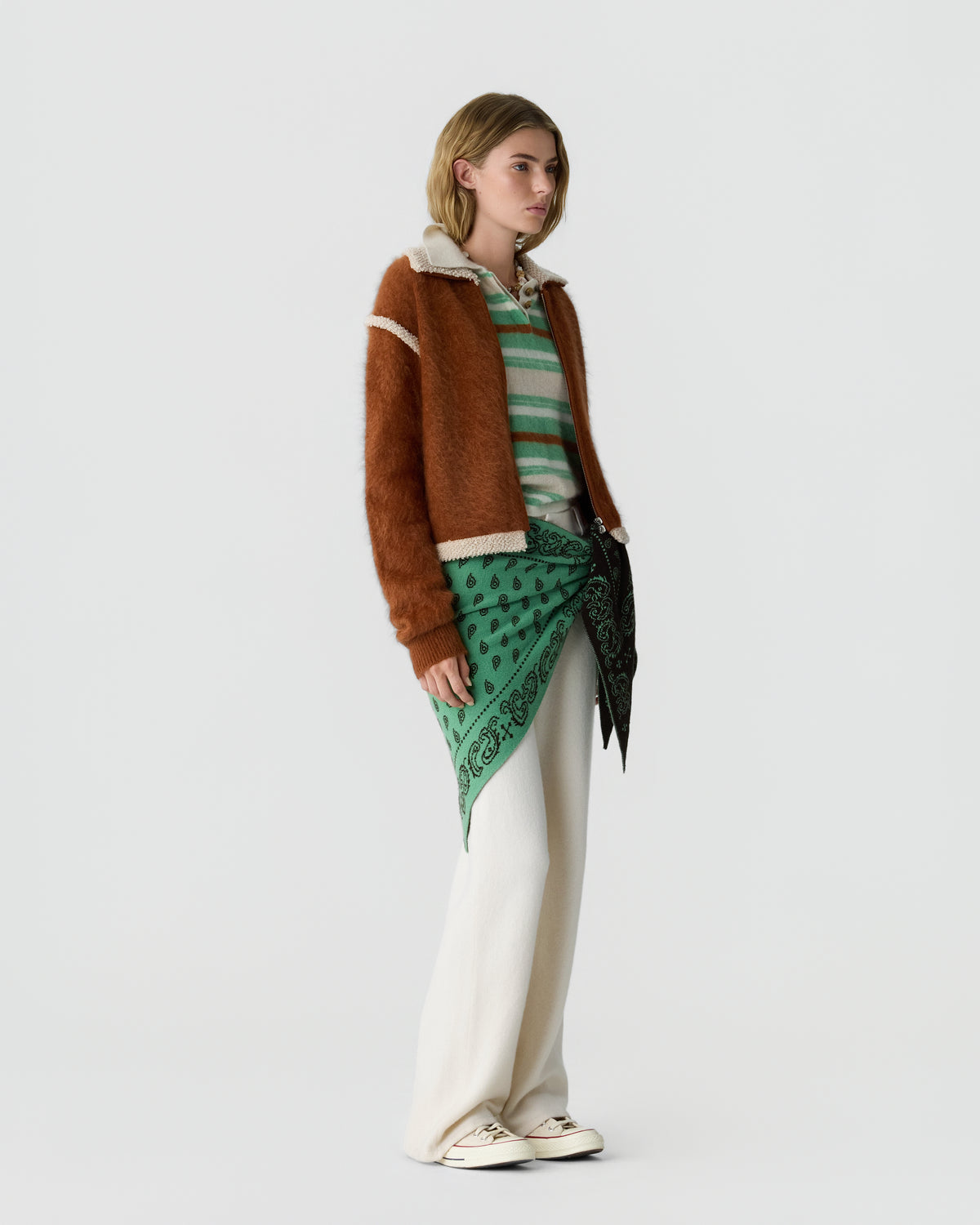 Model wearing a brown cardigan over a green striped top with a patterned green scarf at the waist cream trousers and white sneakers against a light background