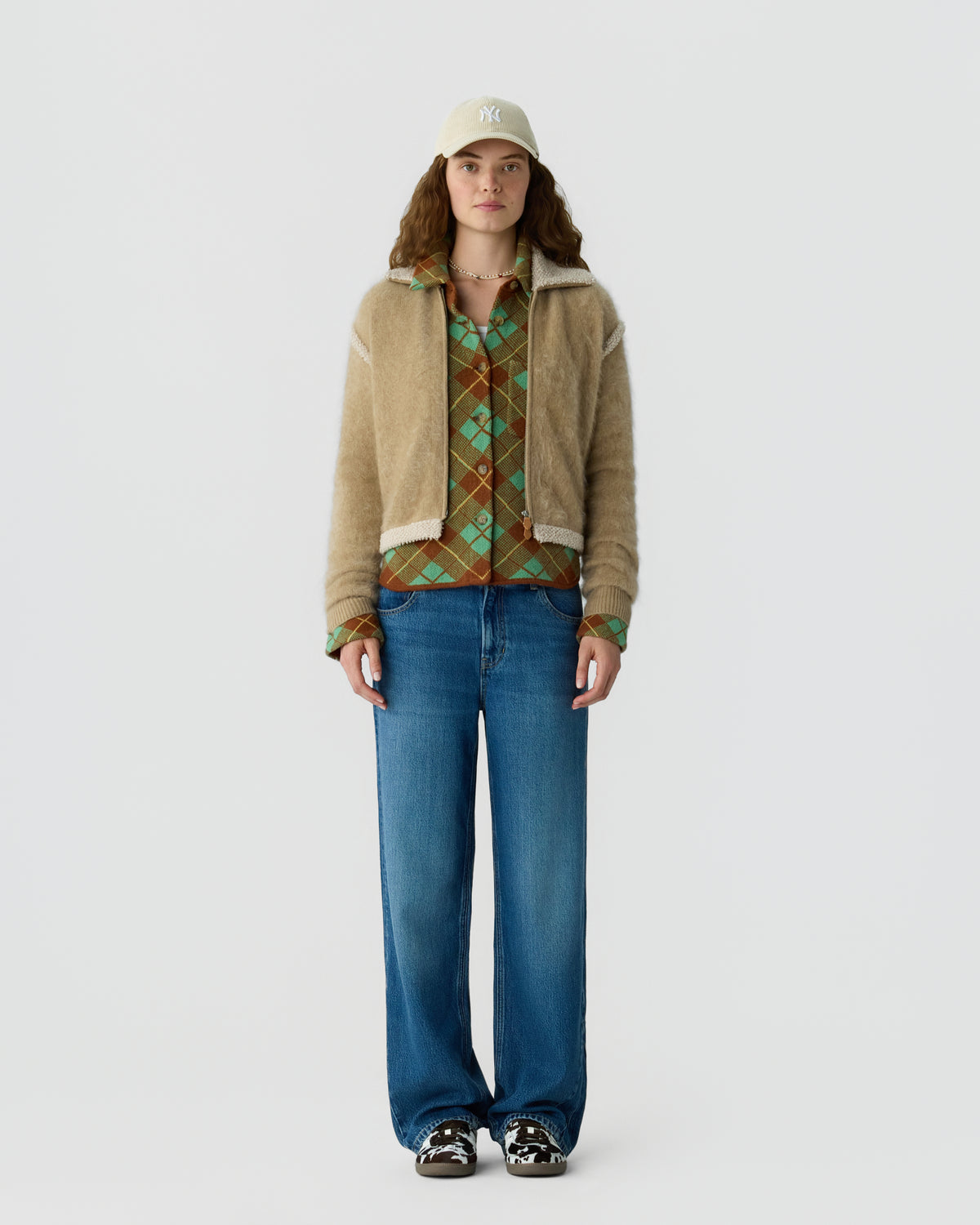 Full length model wearing a beige jacket over a green patterned shirt with blue jeans a light baseball cap and black and white sneakers against a pale background