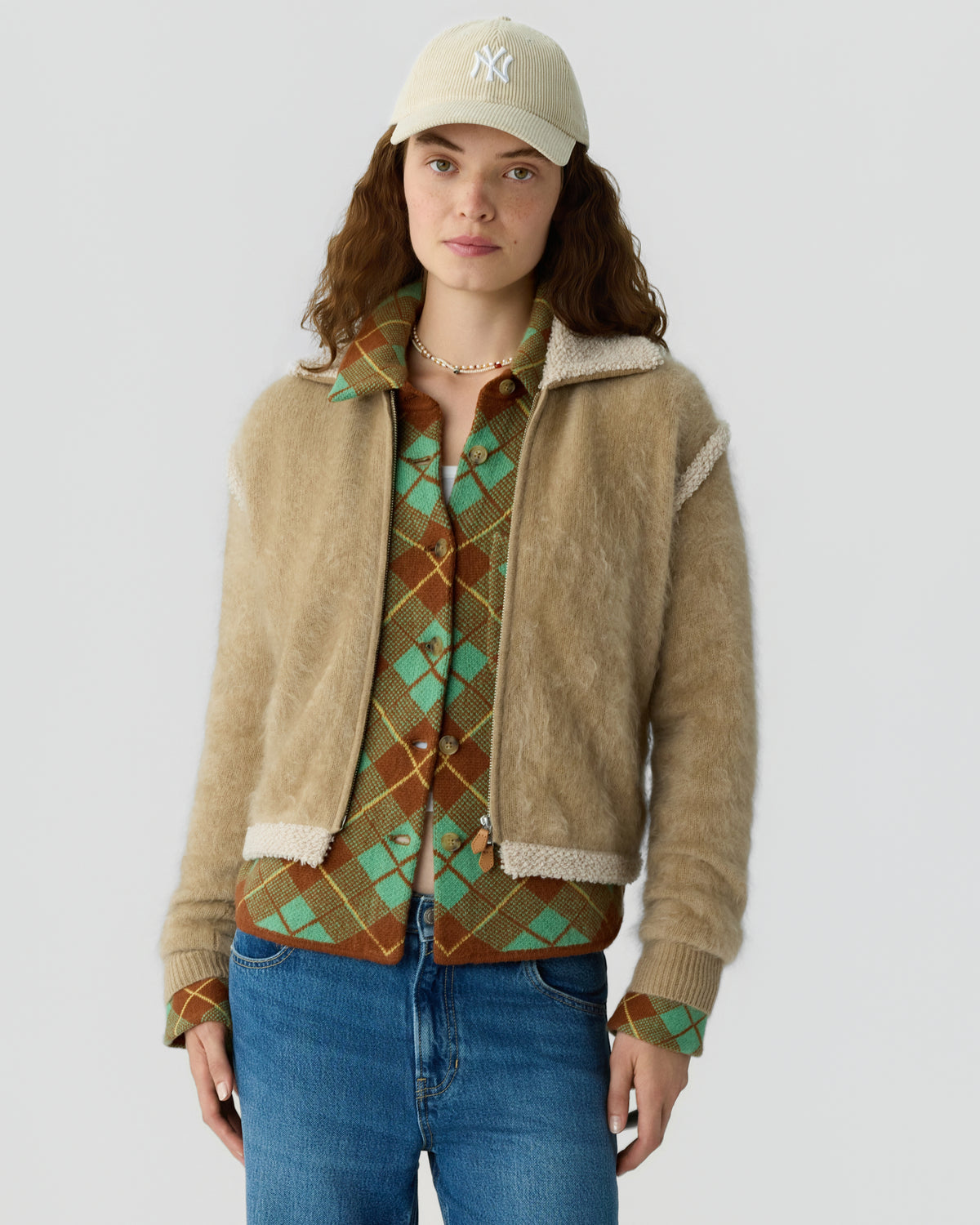 Woman wearing a beige baseball cap, textured tan jacket, patterned green and brown cardigan, and blue jeans against a light gray background