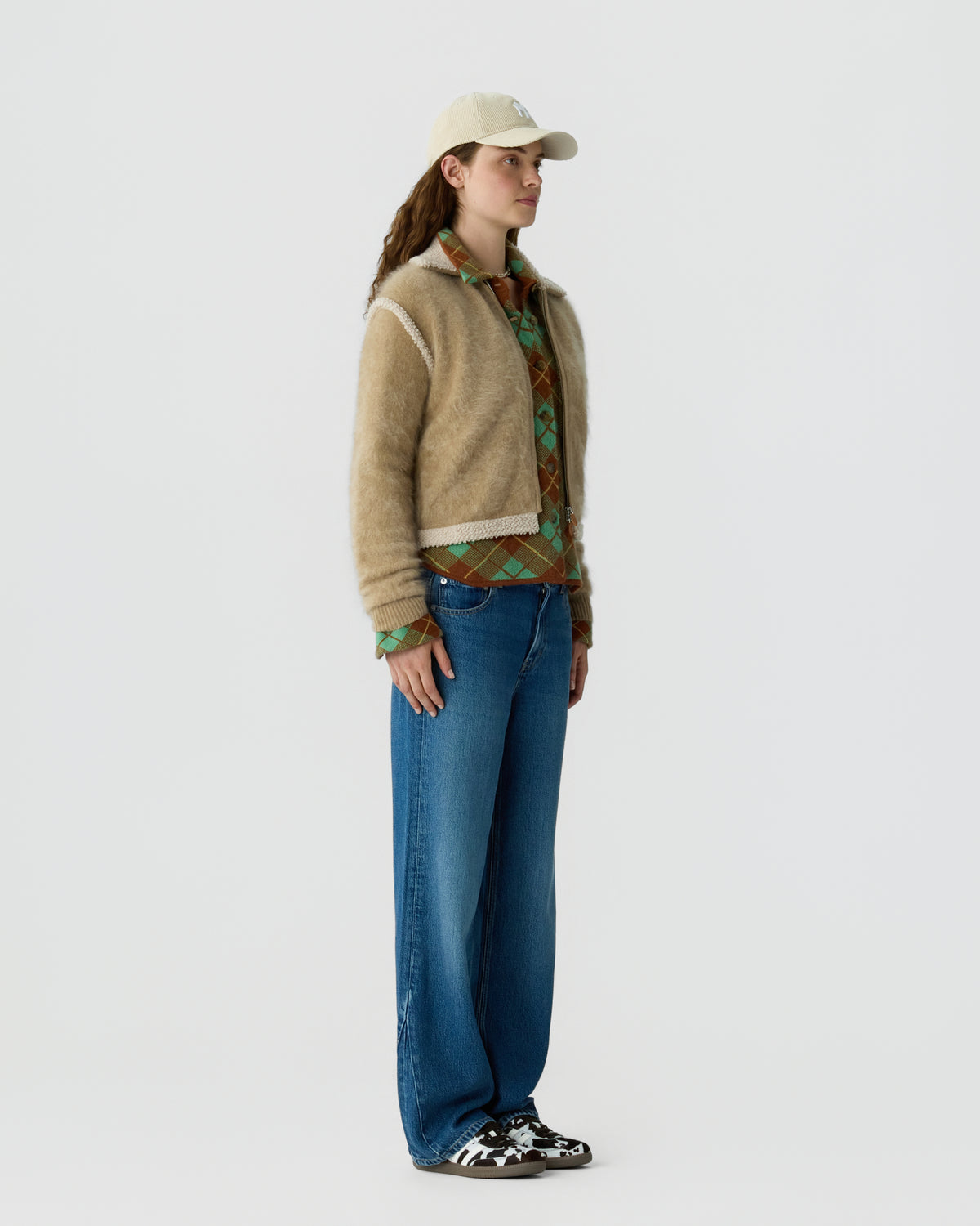 Woman wearing a beige cardigan over a patterned shirt with wide leg blue jeans a light cap and black and white sneakers against a pale background
