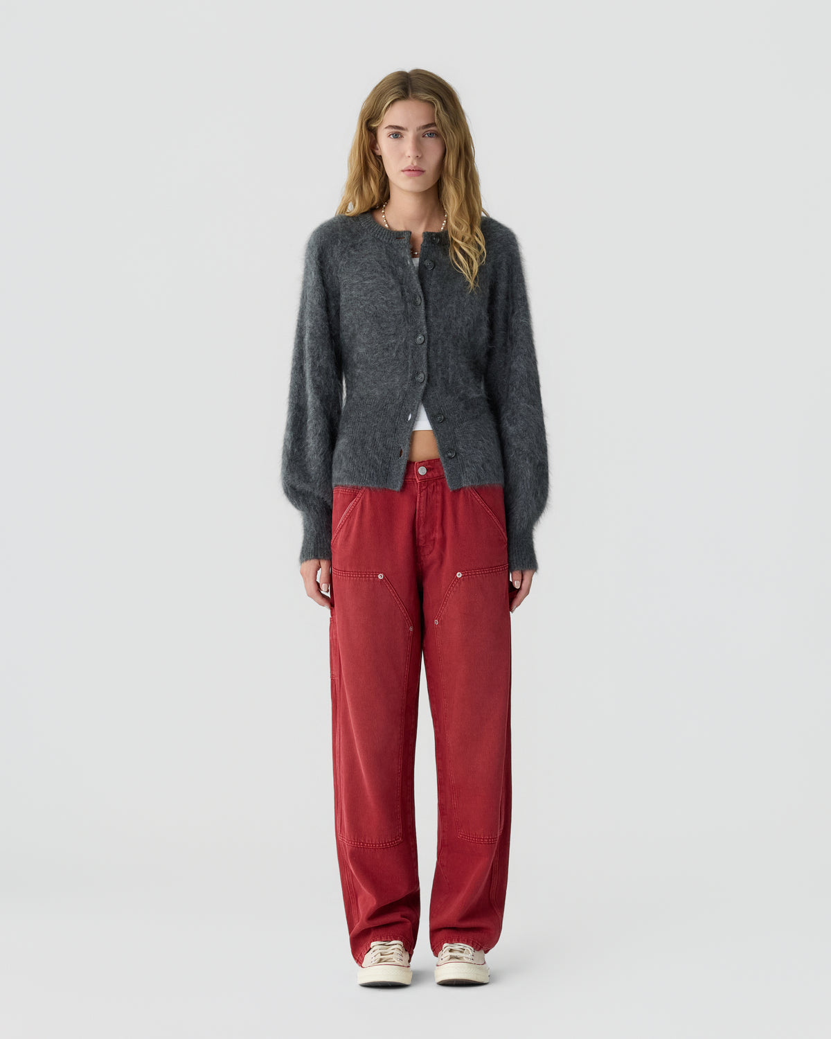 Woman standing against a light gray background, wearing a charcoal buttoned cardigan, red trousers, and cream sneakers.