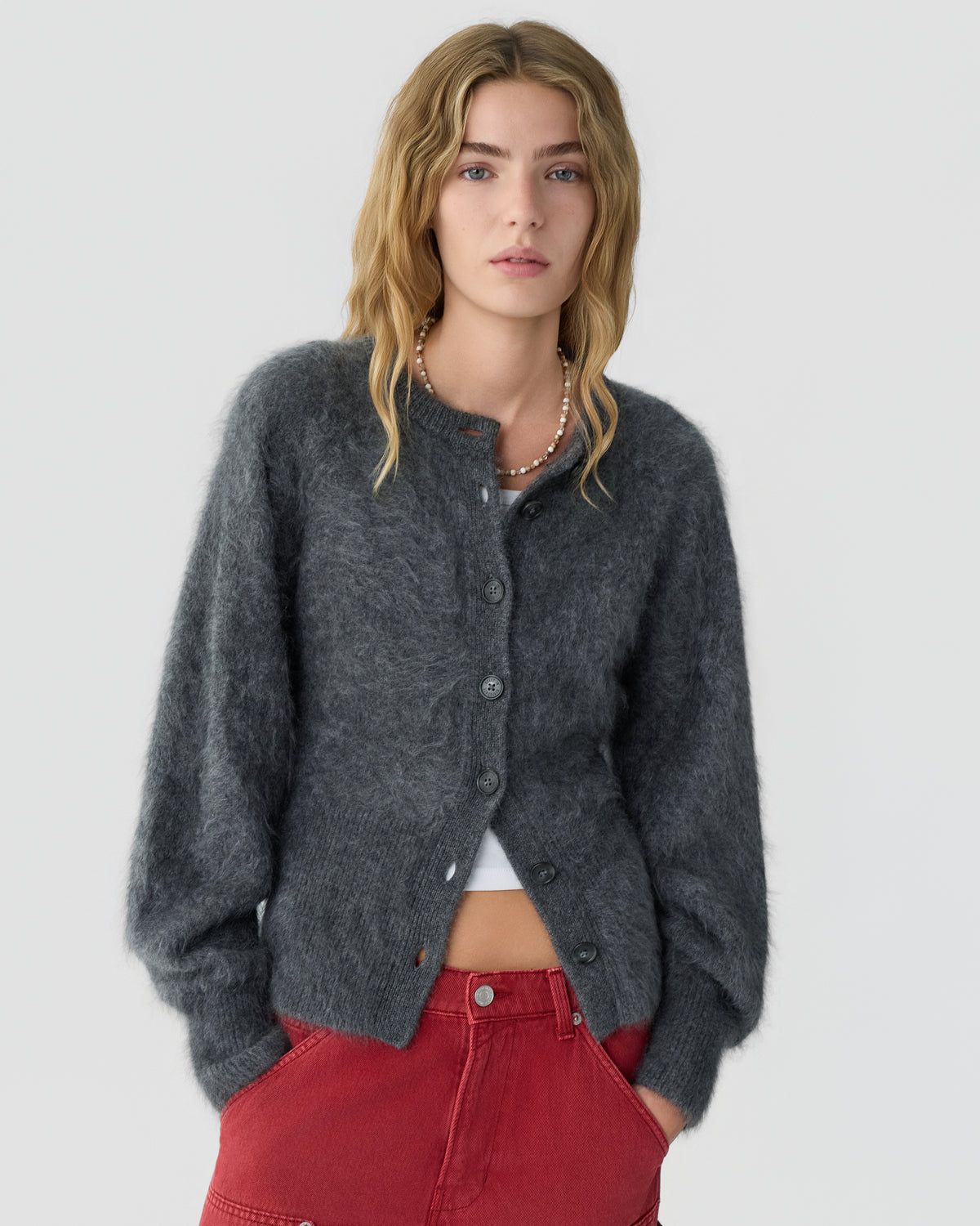 A woman with wavy hair wears a dark gray fuzzy button cardigan over a white top and red high-waisted pants against a light gray background