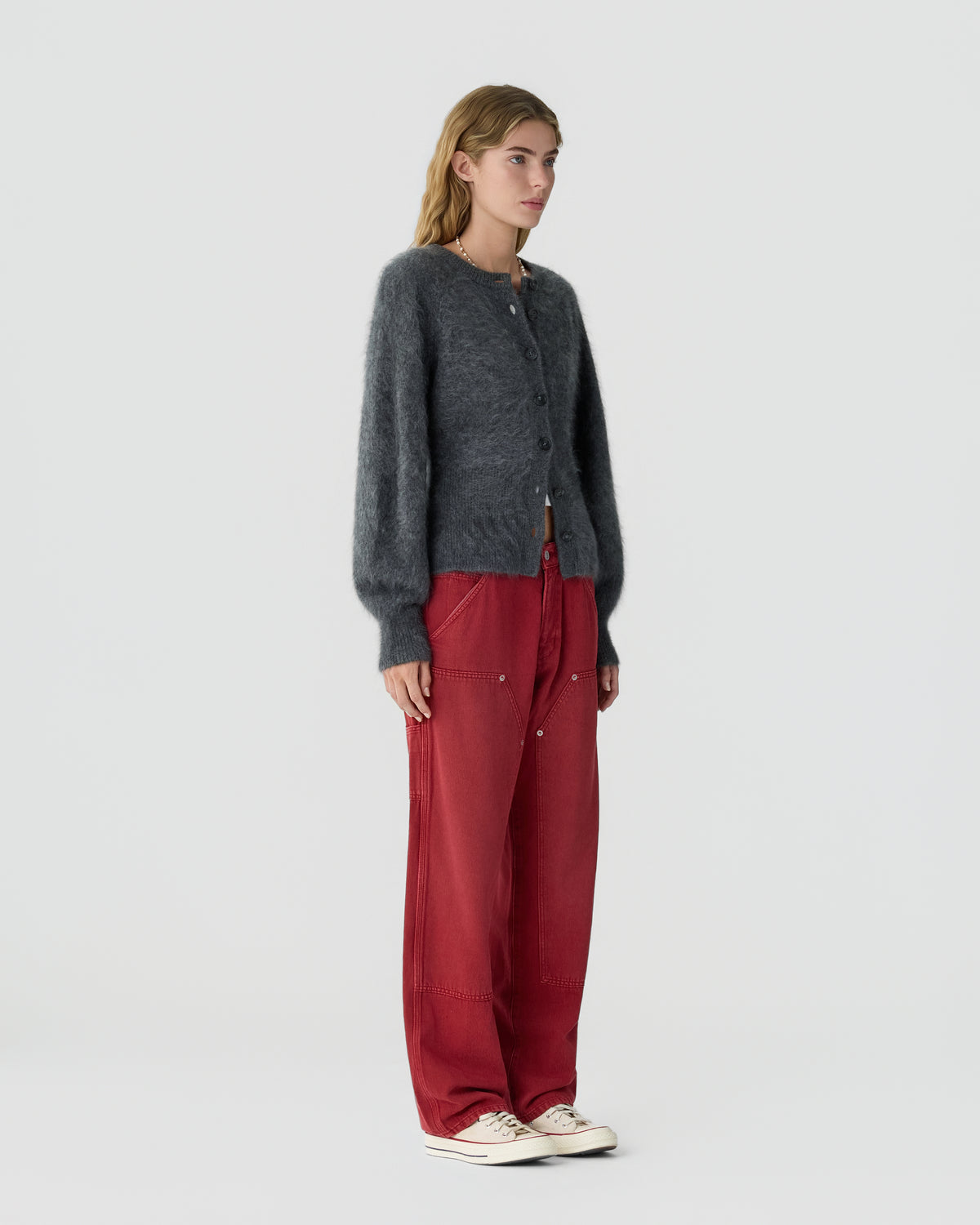 Woman standing in a gray button front cardigan, red trousers, and white sneakers against a plain light background.