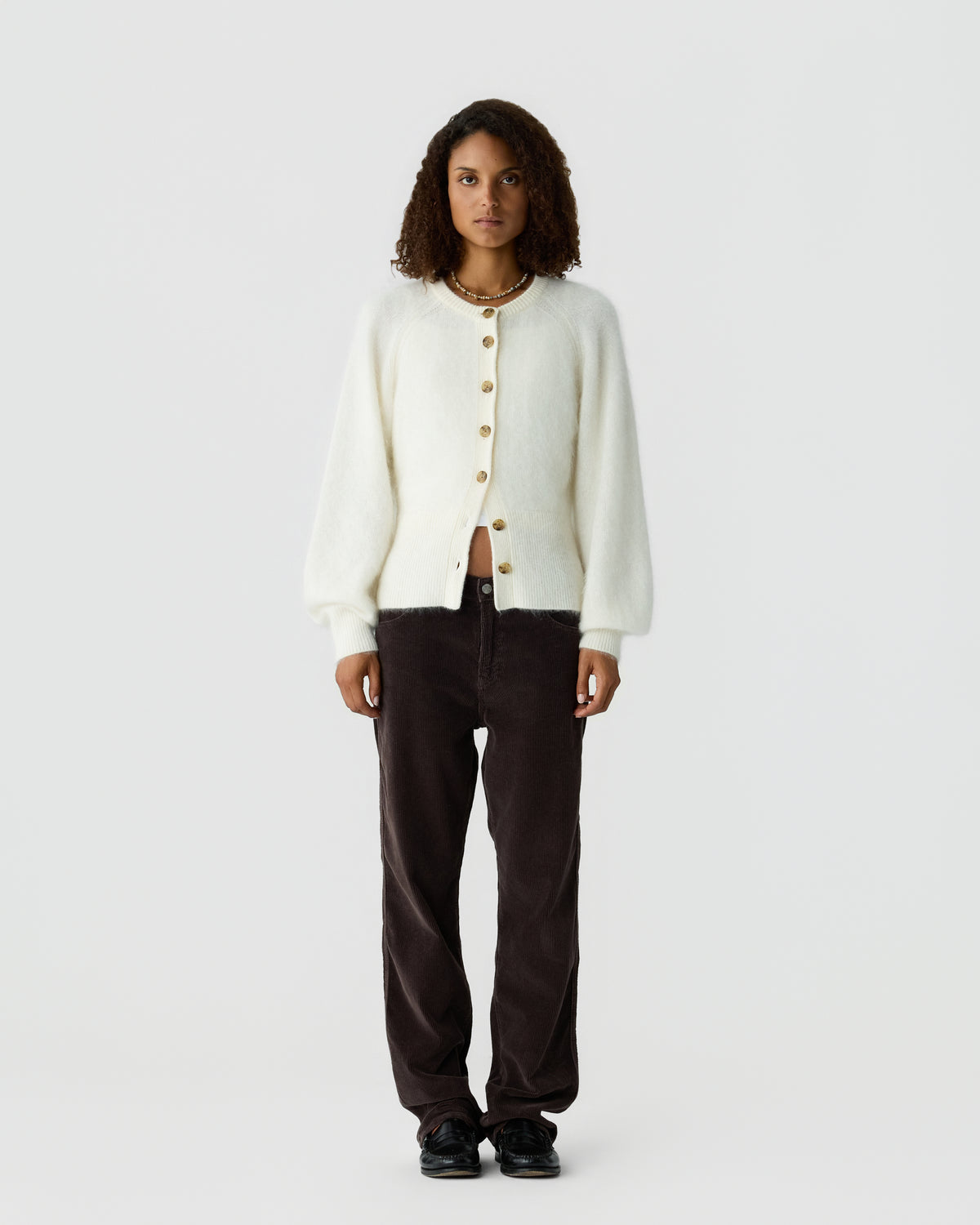 Woman modeling an ivory button front cardigan with dark brown trousers and black shoes against a light gray studio background