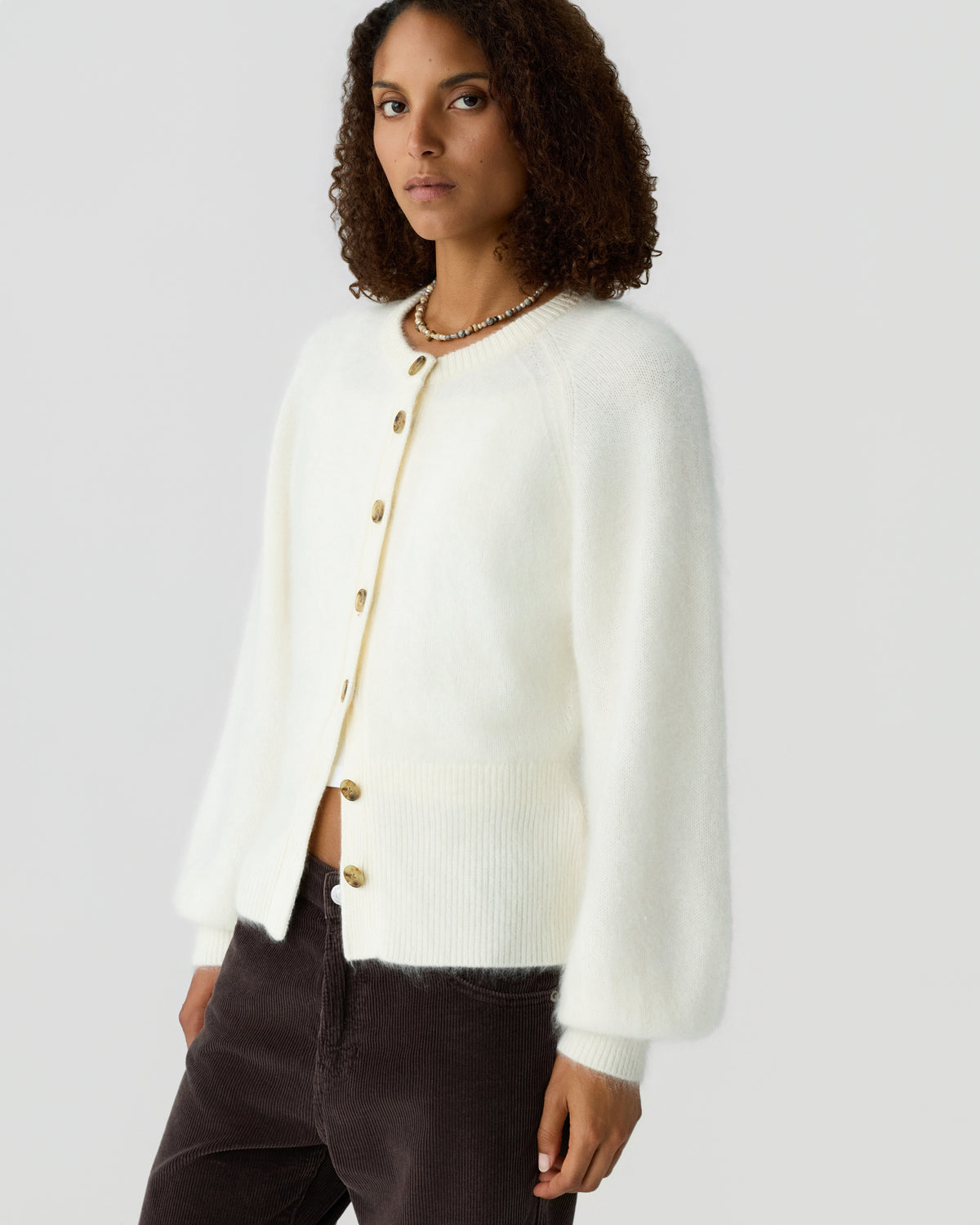 Woman wearing an ivory cashmere cardigan with gold buttons and dark brown trousers against a light gray background.