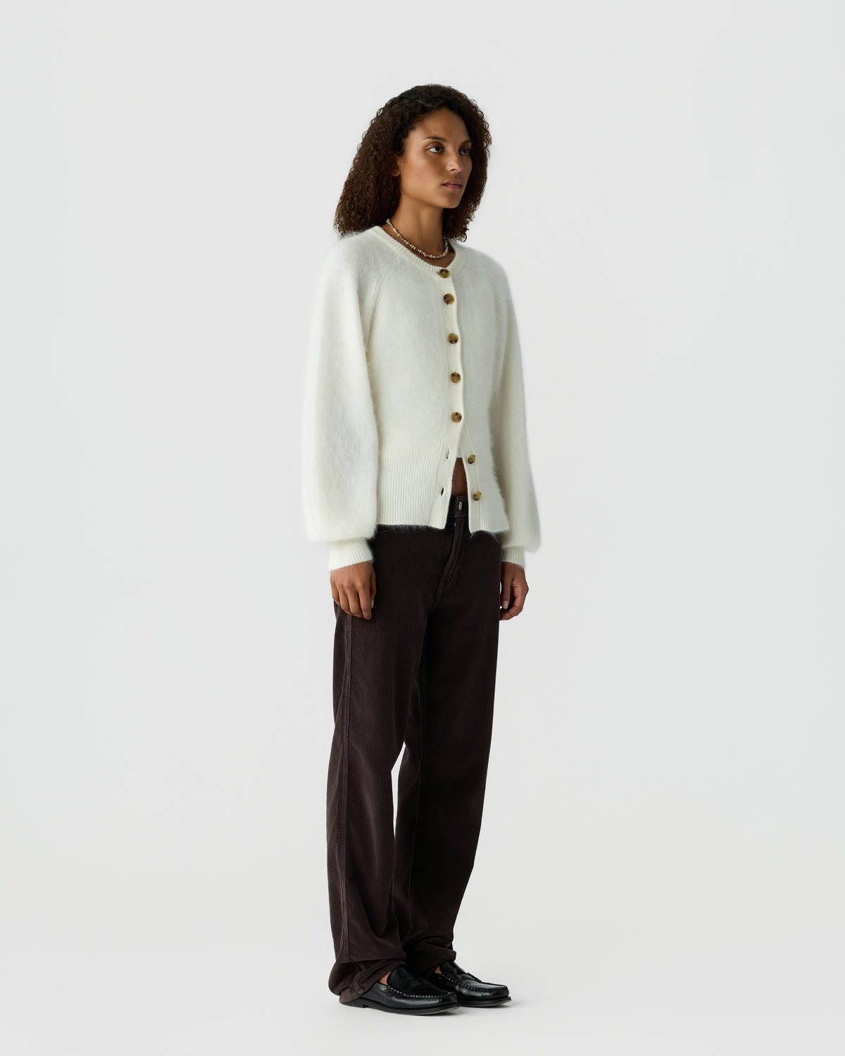 Woman wearing an off white button front cardigan with dark brown trousers and black shoes against a light gray background