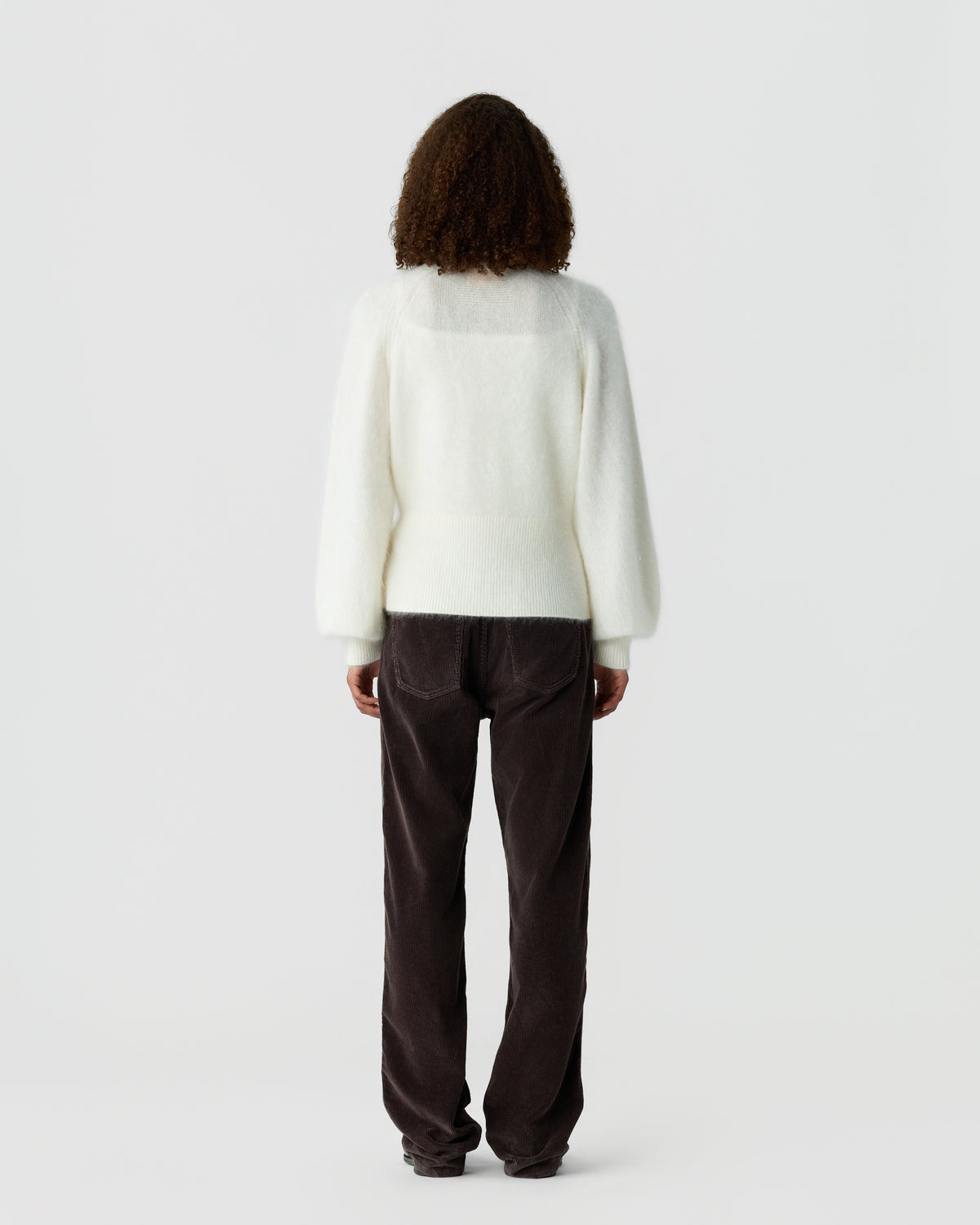 Back view of a person wearing a white long sleeve sweater and dark brown trousers against a plain light gray background