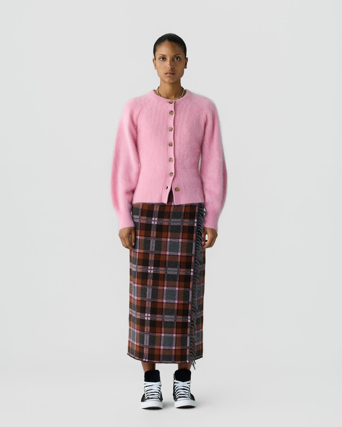 Person standing against a light gray background, wearing a pink buttoned cardigan, long plaid skirt, white striped socks, and black and white sneakers.