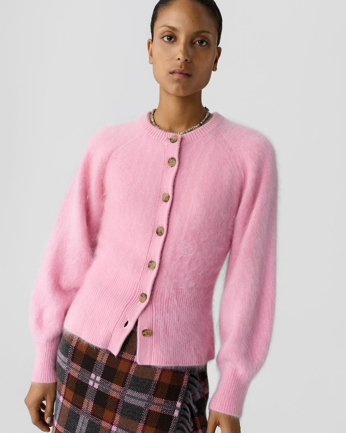 Model wearing a soft pink cashmere cardigan with button front and ribbed cuffs paired with a dark plaid skirt against a light gray background