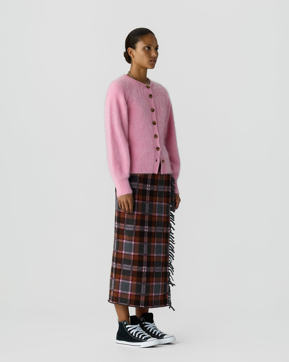 Woman wearing a pink button front cardigan with a plaid midi skirt and black high top sneakers against a light gray background
