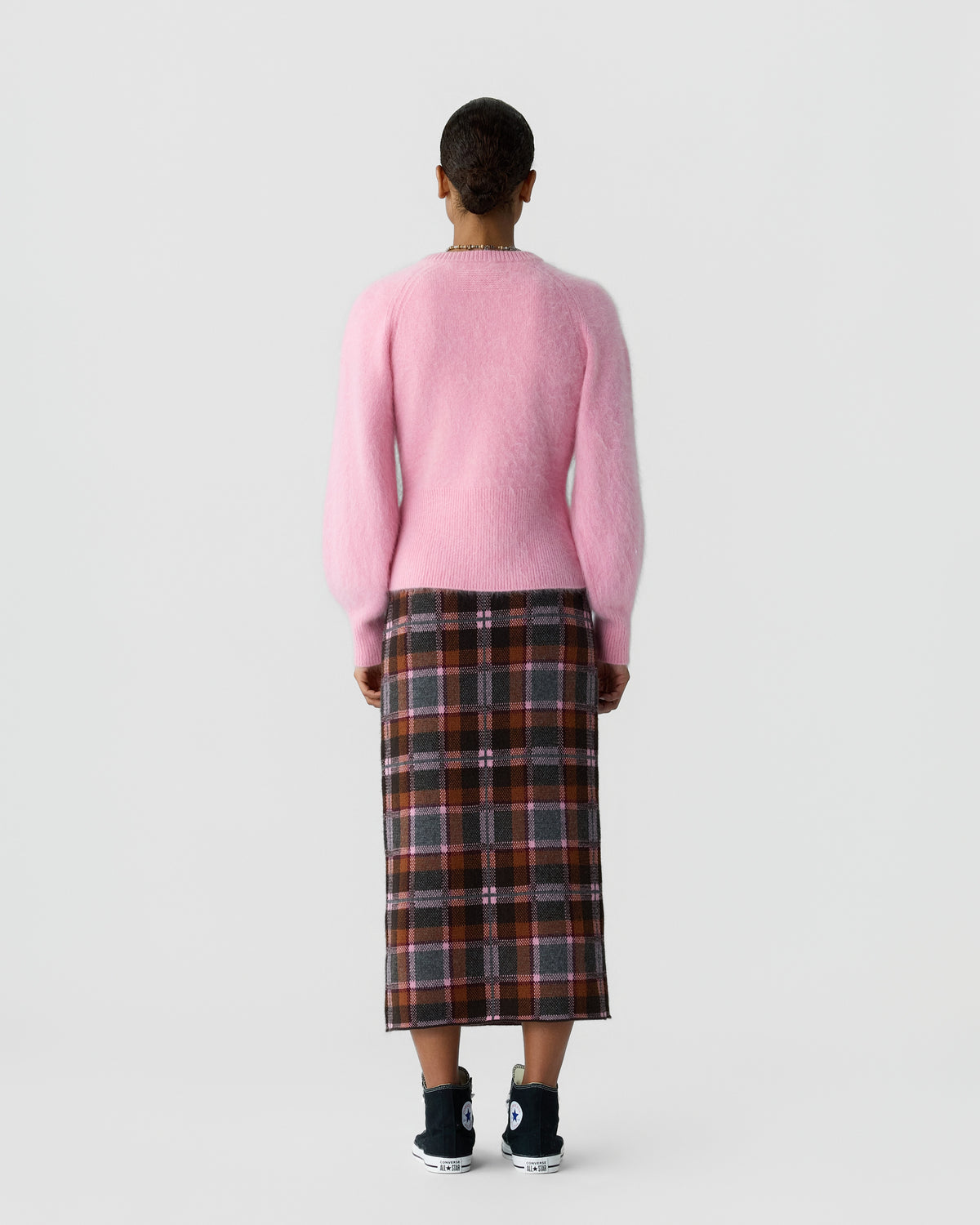 Rear view of a person wearing a pink sweater with a knee length plaid skirt and black sneakers against a light gray background