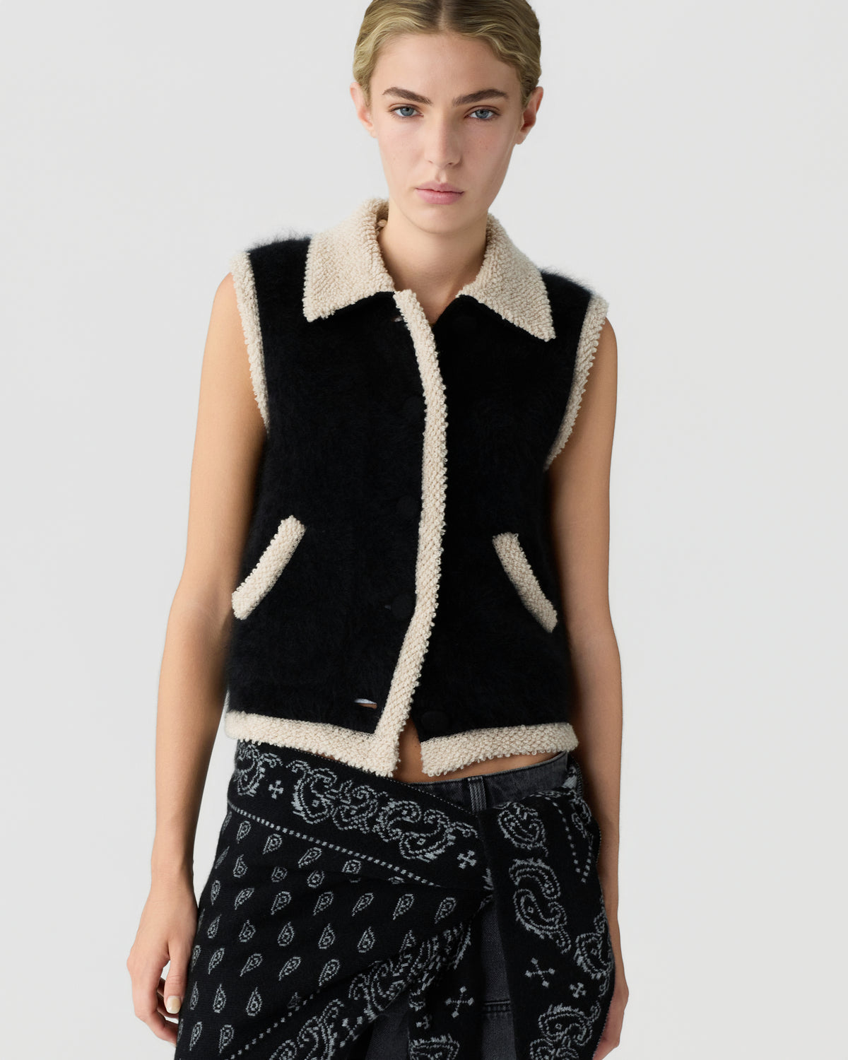 Model wearing a black sleeveless shearling vest with cream trim and collar paired with black patterned trousers against a light gray background