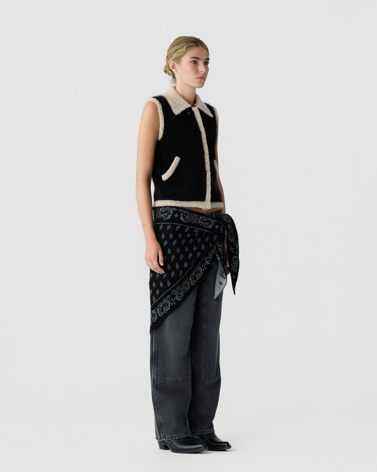 Full length view of a model wearing a black sleeveless knit vest with white trim and pockets over wide leg dark jeans and a patterned scarf at the waist
