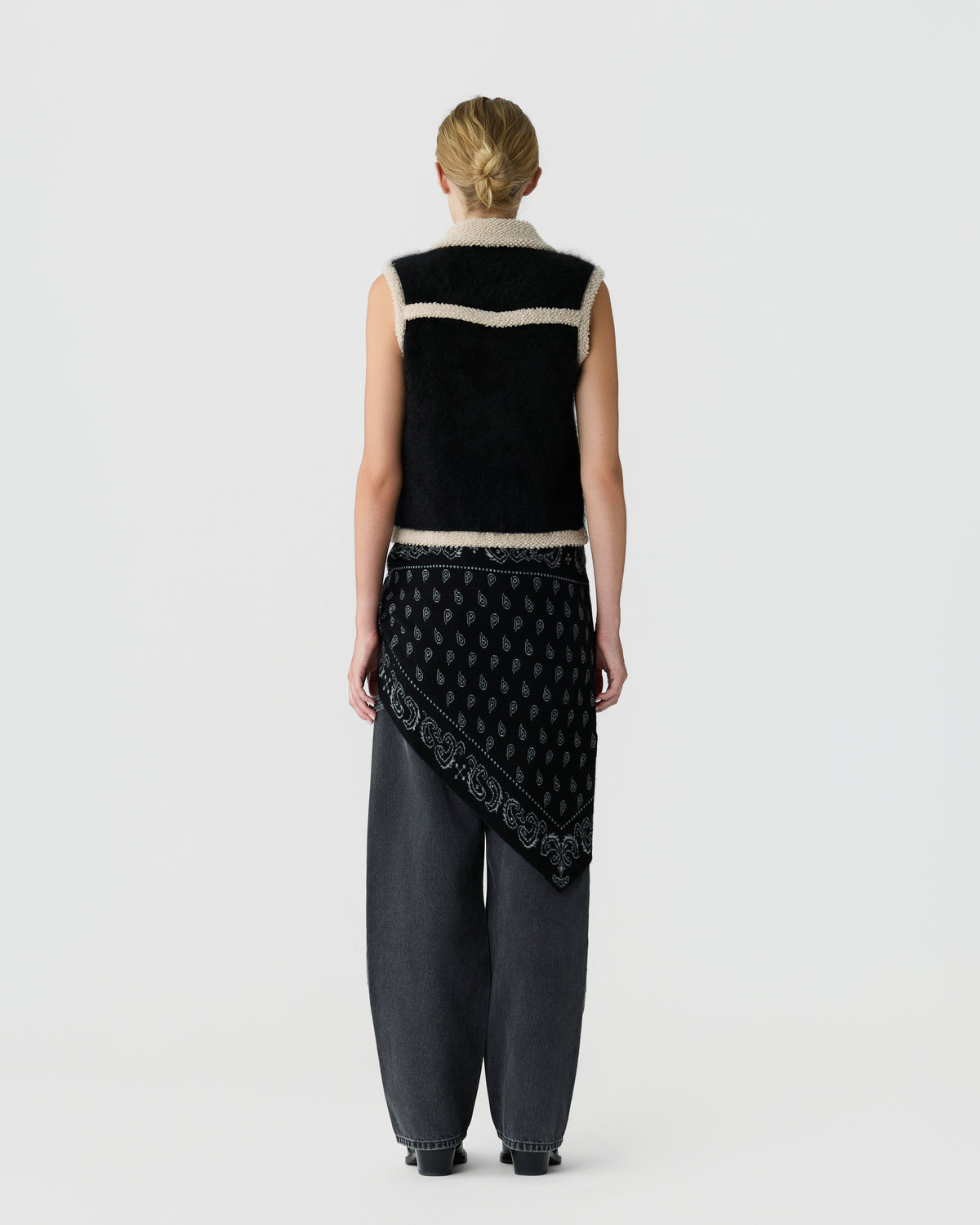 Back view of a woman wearing a black sleeveless knit vest with cream trim and a patterned draped layer over dark relaxed trousers against a light gray background