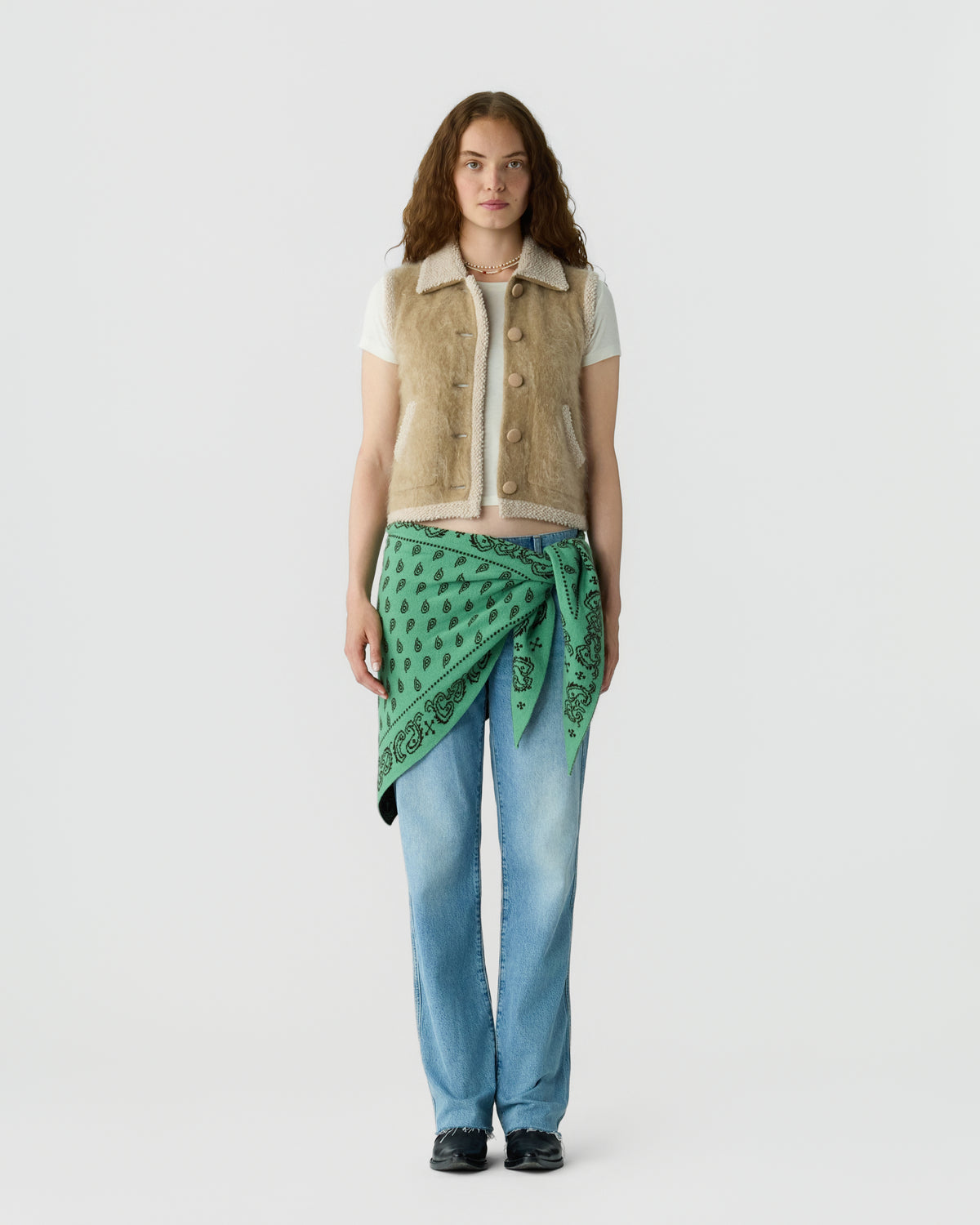 Full length portrait of a woman wearing a cream collared knit vest over a white shirt, blue jeans, a green patterned scarf tied at the waist, and black shoes against a light gray background