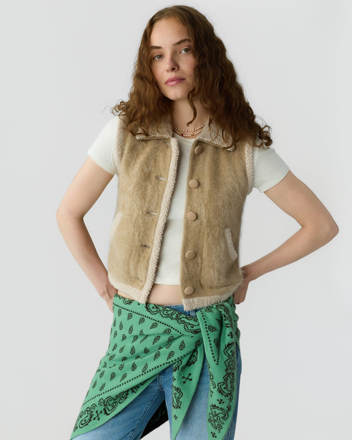 Woman wearing a beige textured button front vest over a white shirt with a green patterned scarf tied at the waist and blue jeans against a light background