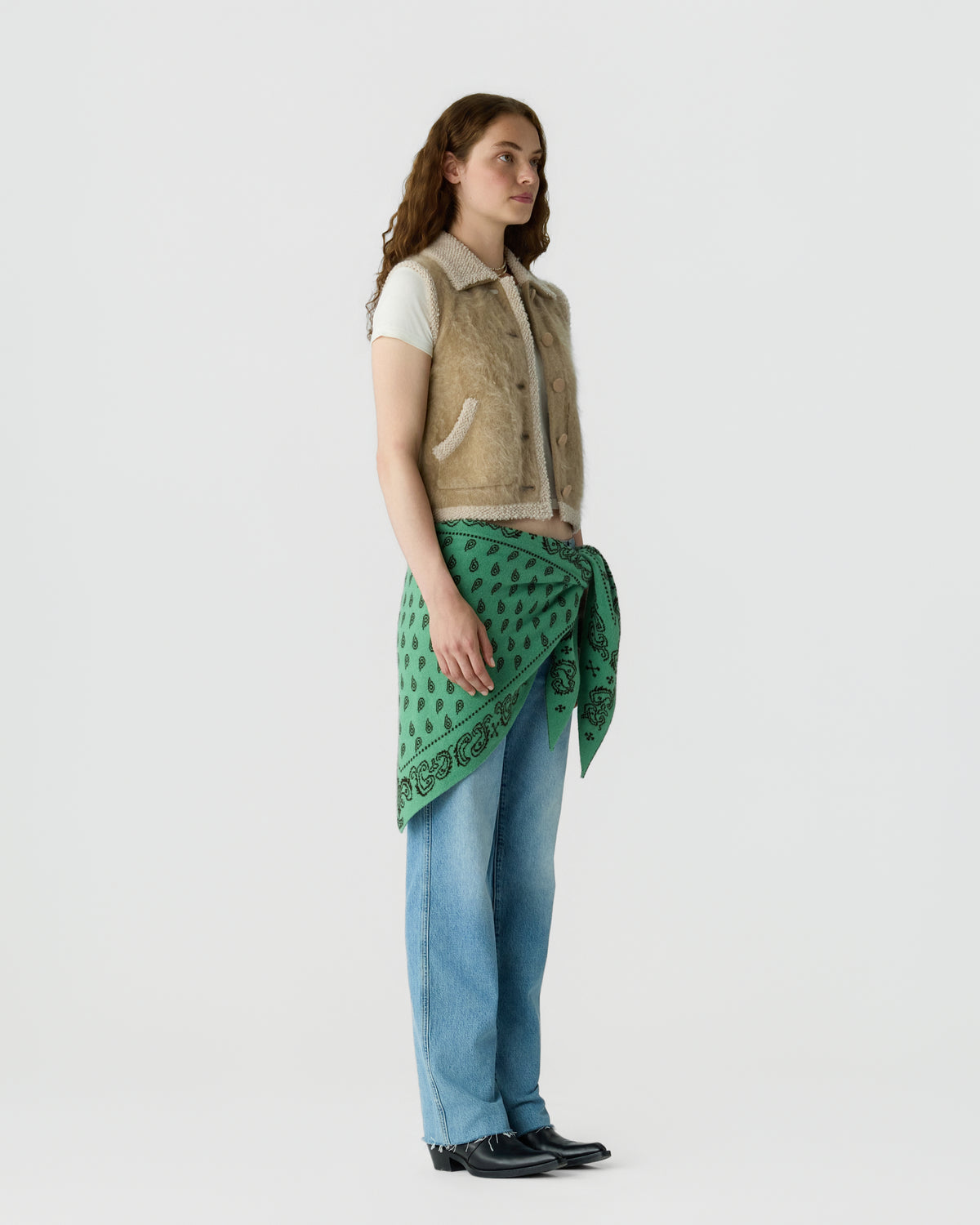 Model wearing a beige sleeveless vest over a white T shirt with a green patterned scarf tied at the waist blue jeans and black loafers against a light gray background