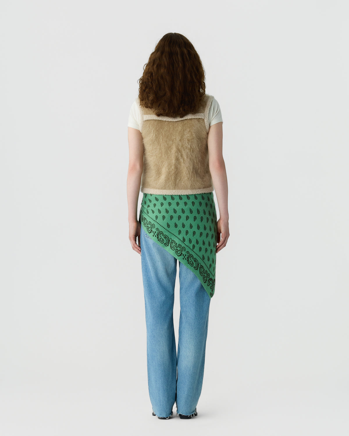 Back view of a woman wearing a cream T shirt, textured beige vest, green patterned scarf tied at the waist, and light blue jeans against a pale background