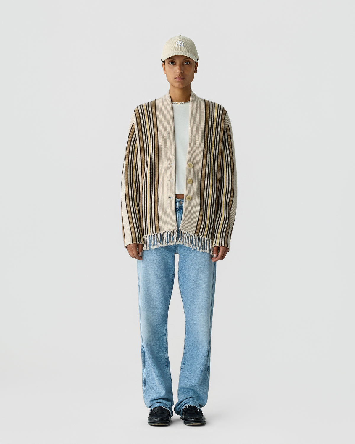 Person wearing a beige and brown vertically striped cardigan with fringe details over a white shirt, light blue jeans, black shoes, and a beige baseball cap standing against a plain light background.