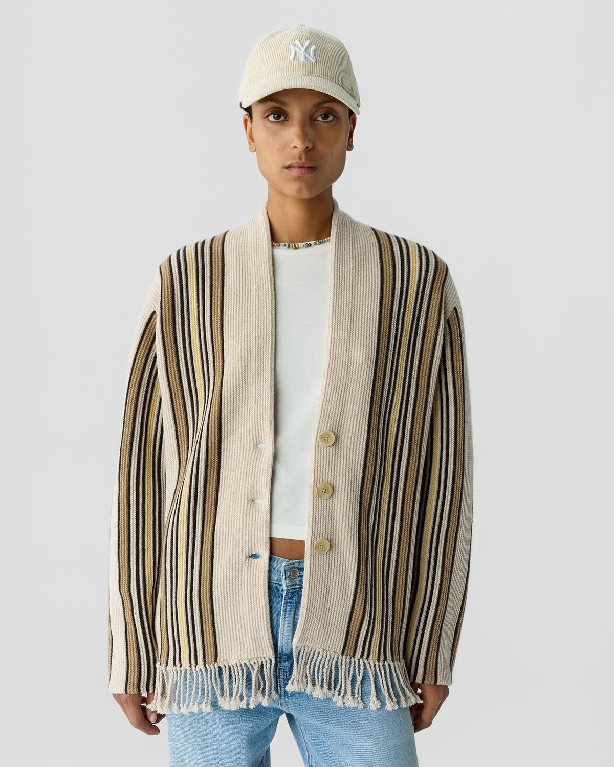 Person wearing a beige, black, and brown striped cardigan with fringe at the hem over a white top and light blue jeans, paired with a beige baseball cap.
