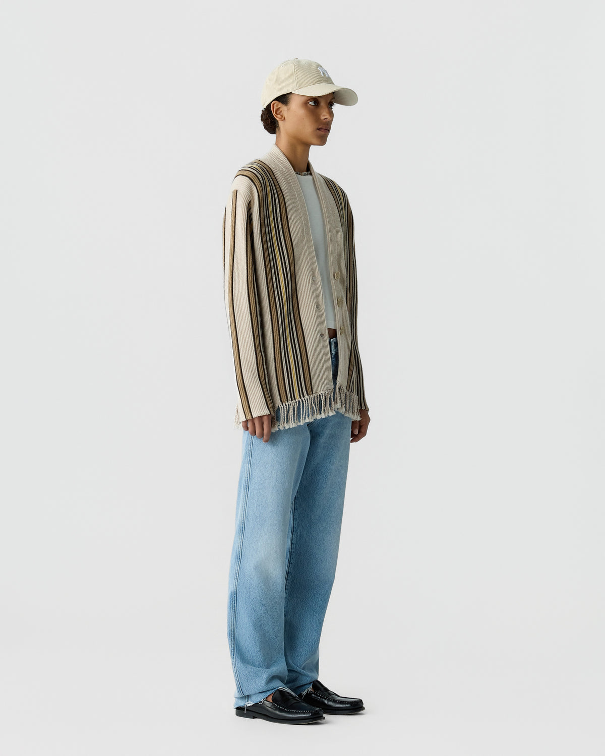 Person standing in profile wearing a beige and brown striped fringed cardigan over a white shirt, light blue wide-leg jeans, black shoes, and a beige cap against a plain white background.