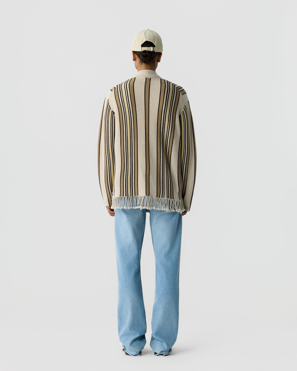 Person wearing a cream and brown vertically striped long-sleeve sweater with fringe at the hem, light blue jeans, a cream baseball cap, standing facing away against a plain background.