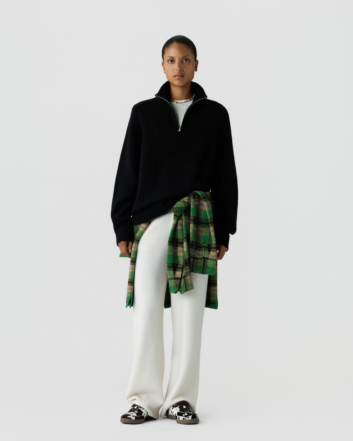 Person wearing a black quarter-zip sweater, white pants, and a green plaid shirt tied around the waist, standing against a plain light gray background.