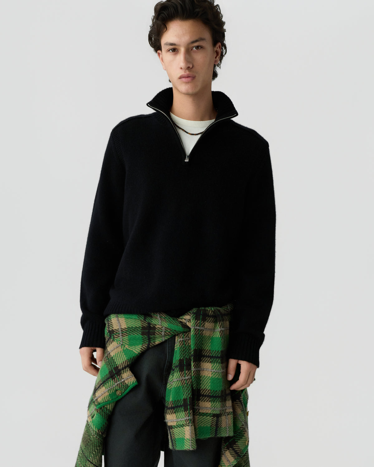 Young man wearing a black quarter-zip sweater layered over a white shirt, with a green and beige plaid shirt tied around his waist against a plain background.