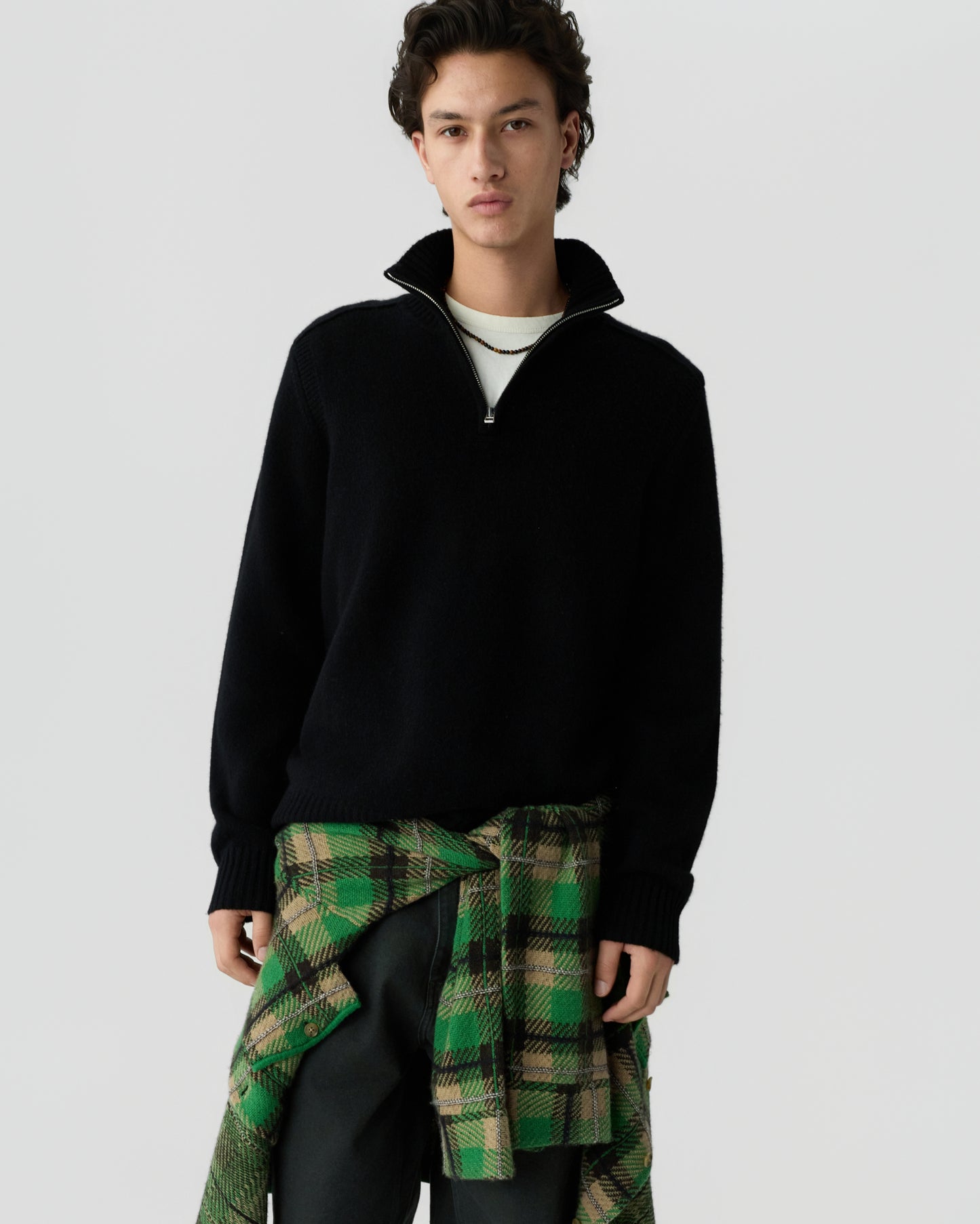 Young man wearing a black quarter-zip sweater layered over a white shirt, with a green and beige plaid shirt tied around his waist against a plain background.