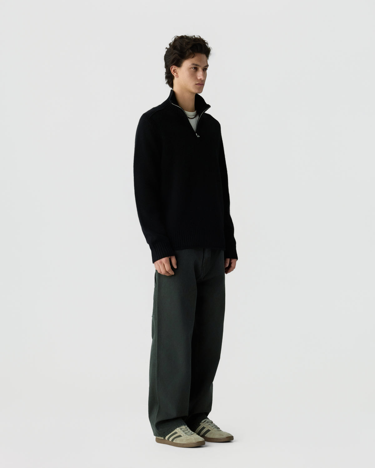 Young man standing against a plain light background wearing a black zip-up sweater, dark wide-leg pants, and beige sneakers with black stripes.