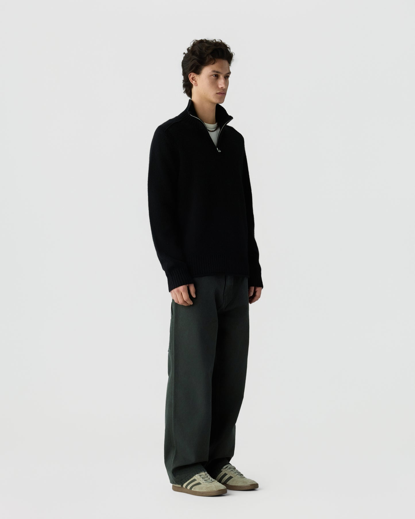 Young man standing against a plain light background wearing a black zip-up sweater, dark wide-leg pants, and beige sneakers with black stripes.