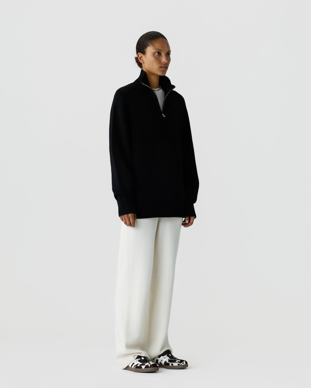 Woman standing against a plain white background wearing a black half-zip sweater, wide-leg white pants, and patterned slip-on shoes.