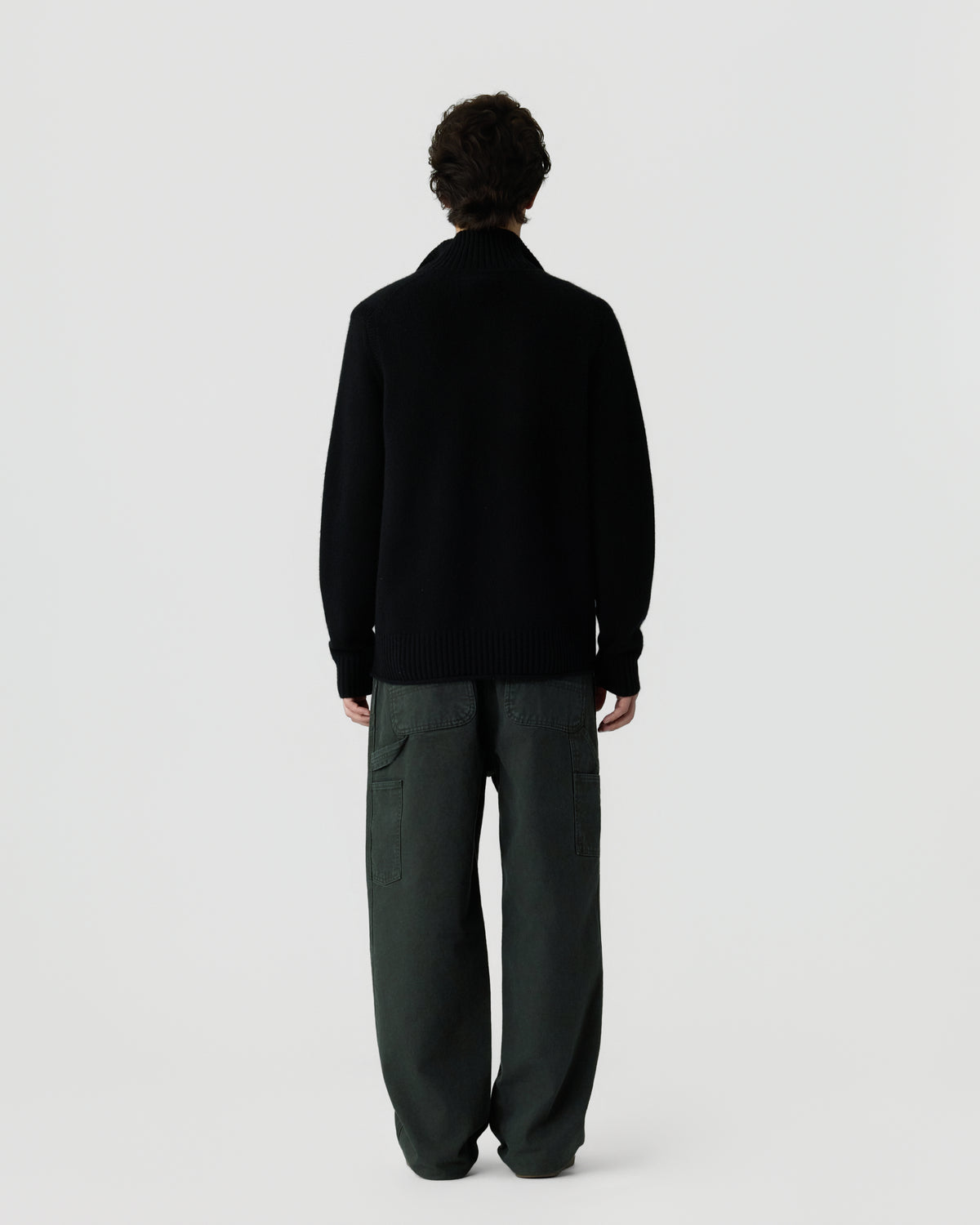 Man standing with back to camera wearing a black cashmere sweater with ribbed collar and cuffs, paired with loose dark green cargo pants against a plain light background.