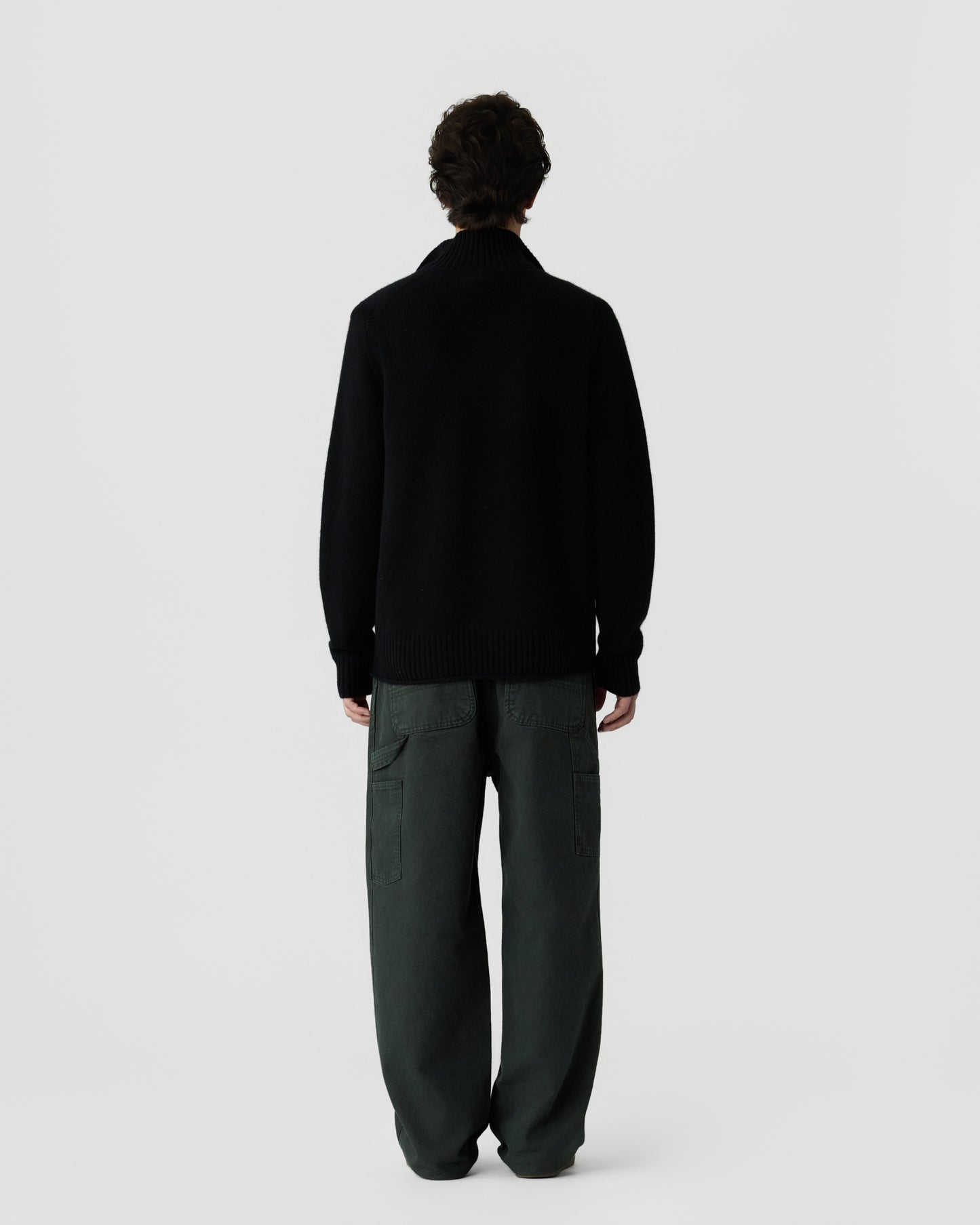 Man standing with back to camera wearing a black cashmere sweater with ribbed collar and cuffs, paired with loose dark green cargo pants against a plain light background.