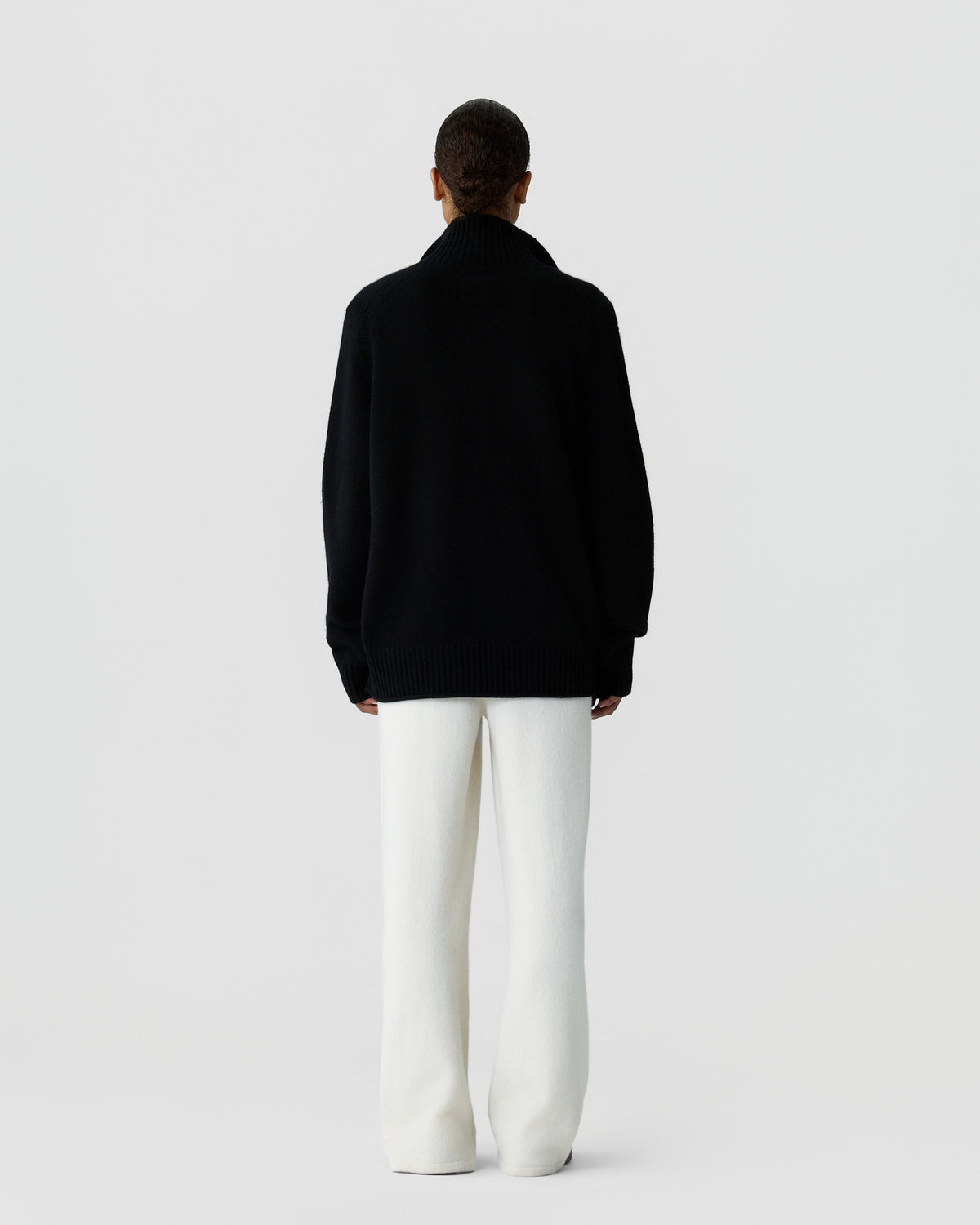 Person seen from behind wearing a loose black turtleneck sweater and wide white pants against a plain light grey background.