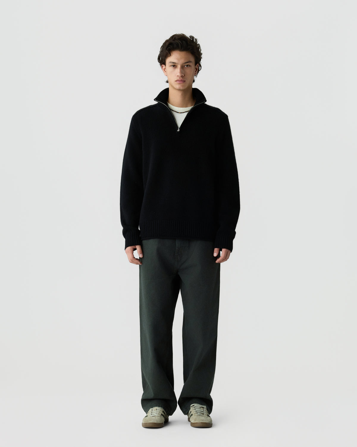 A young man stands against a plain light gray background wearing a black quarter-zip sweater, dark wide-leg pants, and light-colored sneakers.